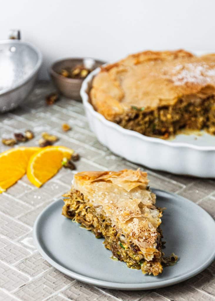 Slice of Moroccan Chicken Pie on a plate. Whole pie with a slice out of it and orange slices in the background.