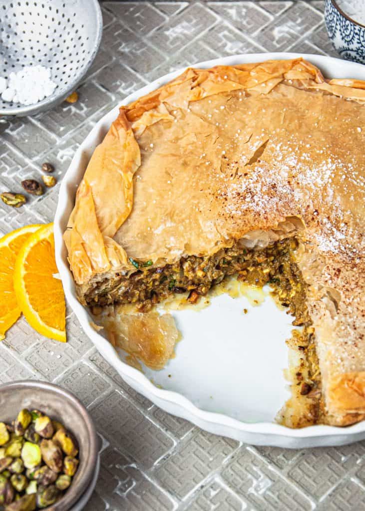 Moroccan Chicken Pie (Bastiya) with a slice out of it, showing inside filling. Bowl of pistachios and orange slices on the side.