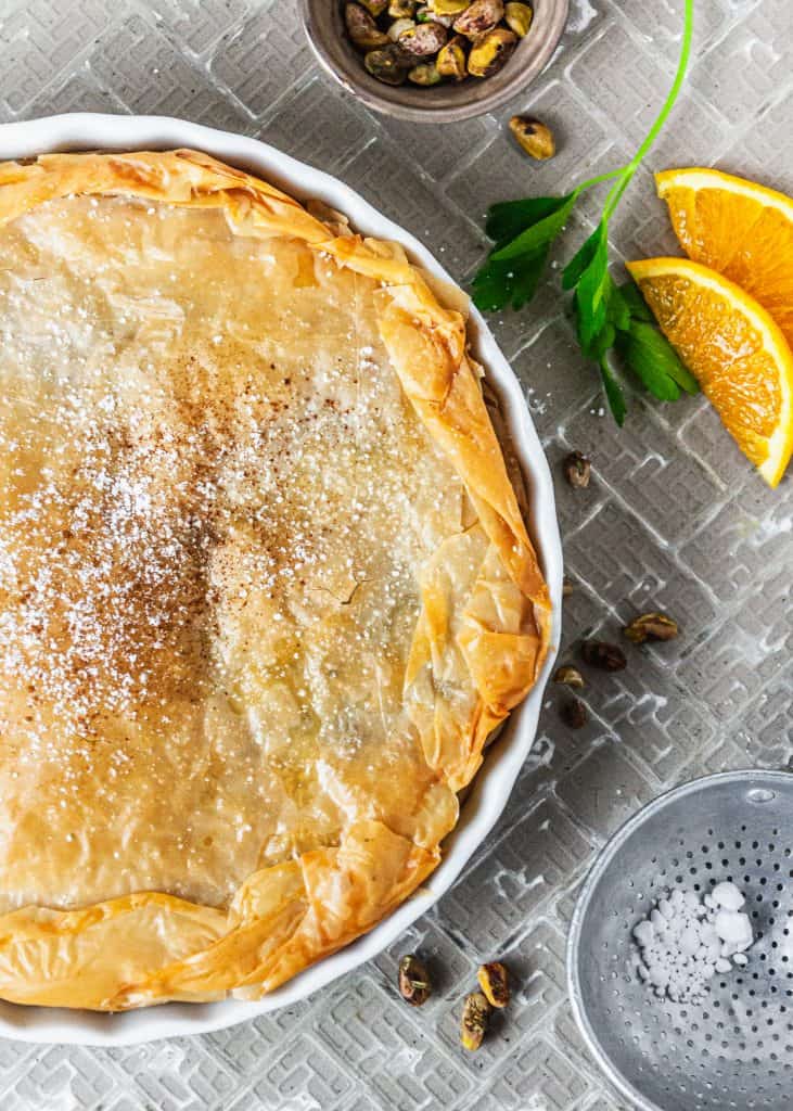 Moroccan Chicken Pie (Bastiya) overhead view, half of pie in left side of frame, on gray tiled surface. Surrounded by pistachios, orange slices, parsley sprigs. 