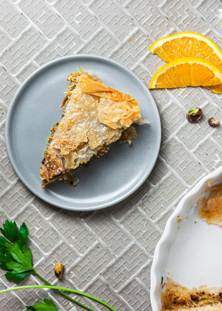 Slice of Moroccan Chicken Pie on a plate, overhead view showing flaky phyllo dough crust. Two orange slices, some pistachios, and parsley sprigs to the side of the plate.