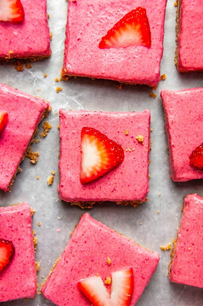 Square cut strawberry dessert bars on a sheet tray seen from overhead with scattered graham cracker crumbs around. Each square is topped with a strawberry slice.