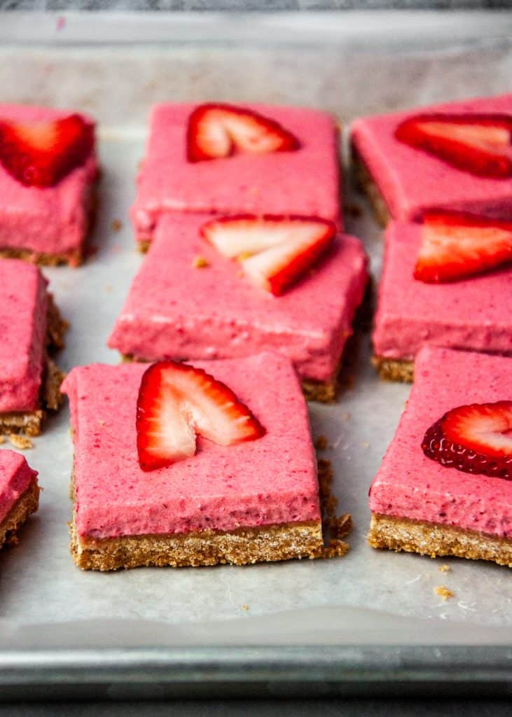 Square cut strawberry dessert bars on a sheet tray with scattered graham cracker crumbs. Front row of bars in focus, others blurry in background.