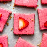 square cut strawberry dessert bars on a sheet pan, each slice topped with a strawberry slice, scattered crumbs around