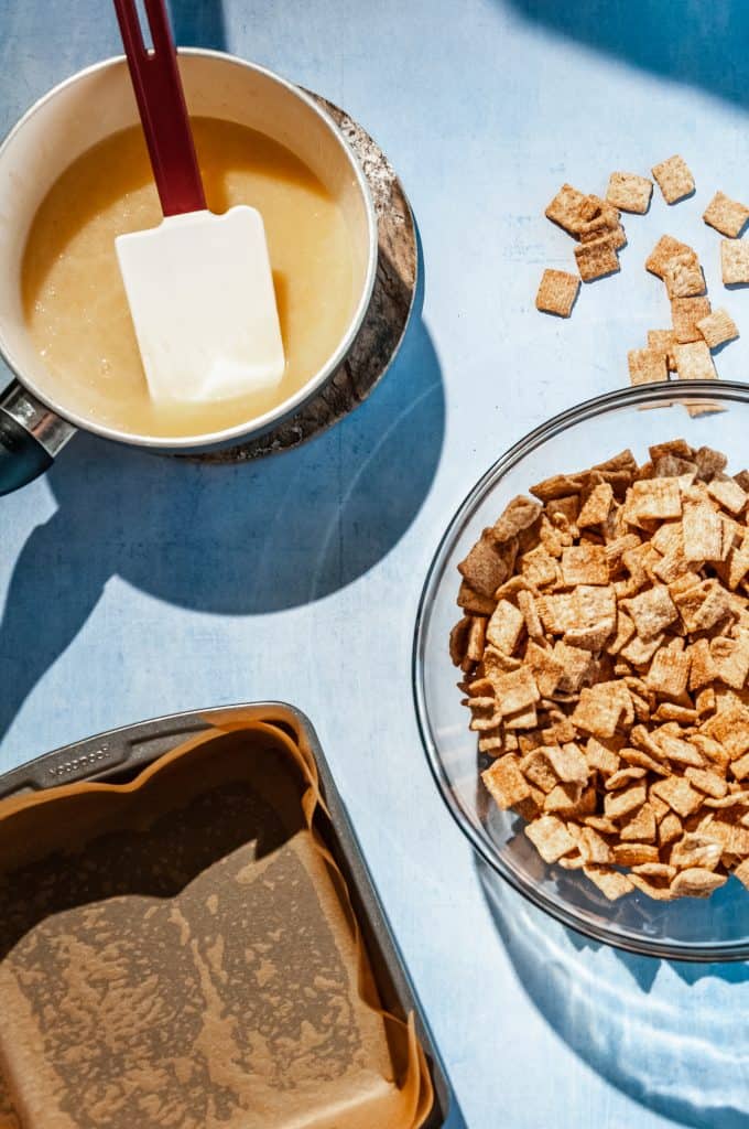getting ready to make homemade milk and cereal bars. cereal in large bowl, baking pan lined with parchment paper, syrup mixture in a saucepan