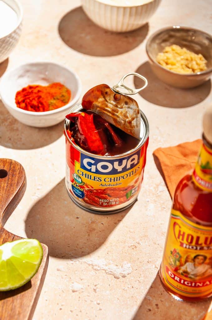 Open can of Goya chipotle chiles in adobo sauce, surrounded by other ingredients needed to make chipotle sauce: lime, hot sauce, spices, garlic.