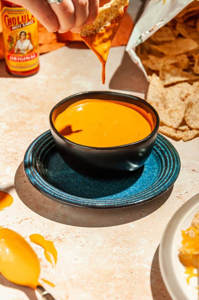 A hand dipping a tortilla chip into a bowl of chipotle dipping sauce, chipotle sauce dripping off of the chip. Bag of tortilla chips and bottle of hot sauce in the background. 