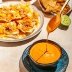 Chipotle sauce dripping off a spoon into bowl filled with chipotle sauce, a plate of nachos and a bag of tortilla chips in the background.