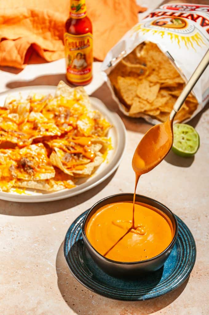 Chipotle sauce dripping off a spoon into bowl filled with chipotle sauce, a plate of nachos and a bag of tortilla chips in the background.