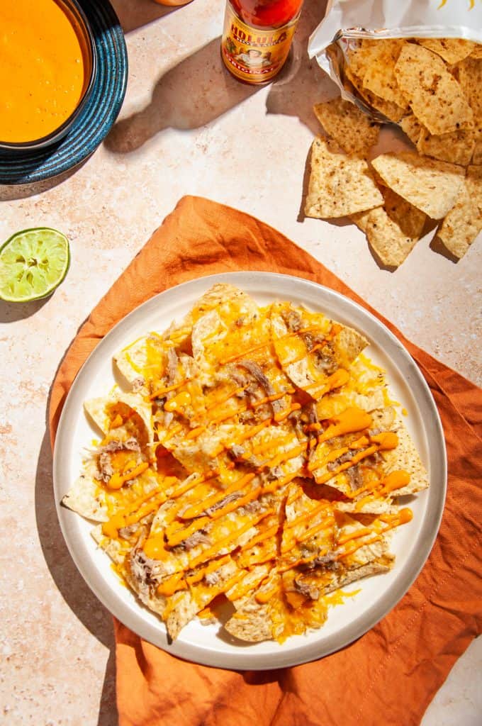 A plate of nachos topped with cheese, pulled pork, and a drizzle of chipotle lime sauce on an orange linen. Extra chipotle sauce, a lime wedge, and tortilla chips to the side.