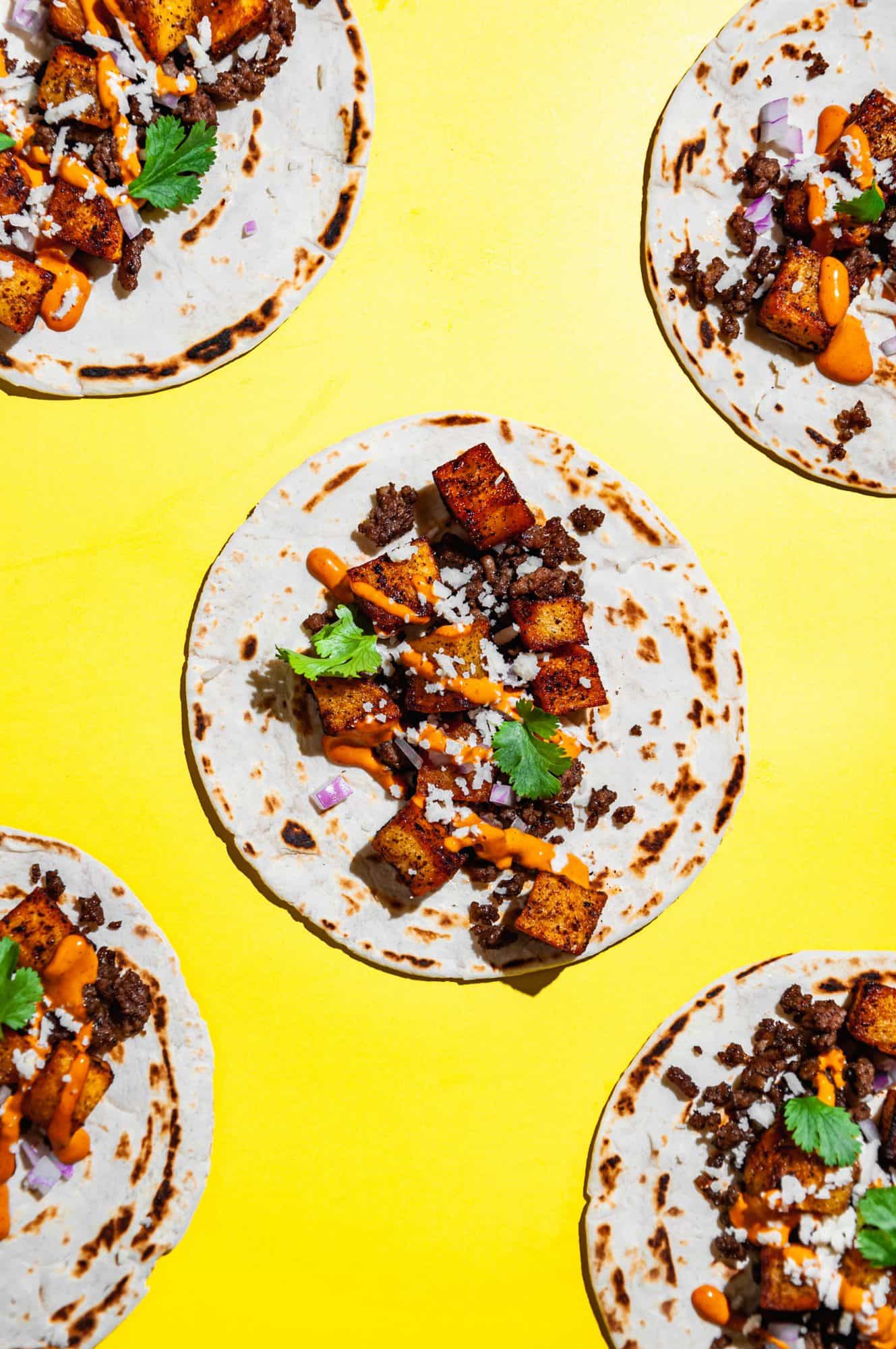 Close up overhead shot of a crispy potato soft taco with ground beef and other garnishes on a bright yellow background. Taco is framed by 4 other potato tacos in 4 corners of the frame. 