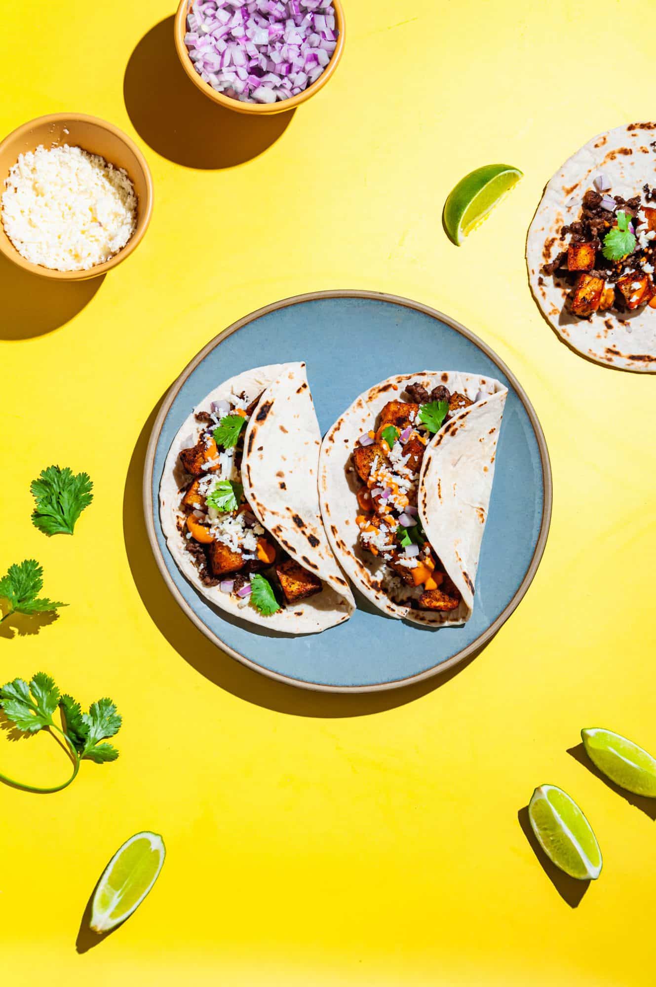 Two crispy potato soft tacos with ground beef on a blue plate, surrounded by a bowl of queso fresco, lime wedges, cilantro, and another taco in top right corner. 