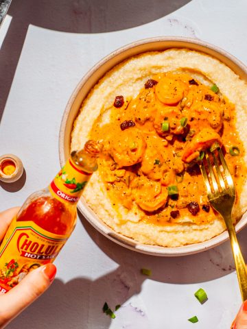 hands holding hot sauce and a fork above a shallow bowl with shrimp in a cajun cream sauce over grits