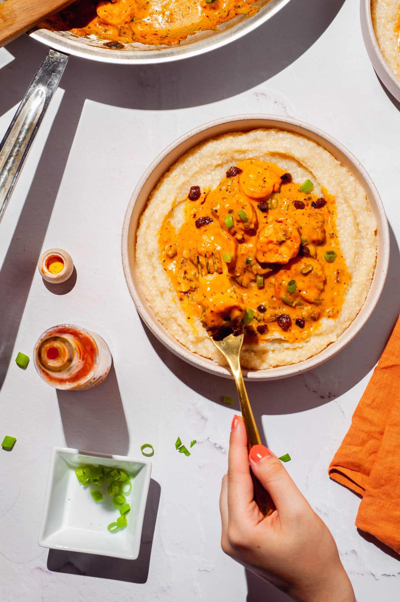 hand holding fork with shrimp over bowl of shrimp and grits with cajun sauce, hot sauce and scallions to the side