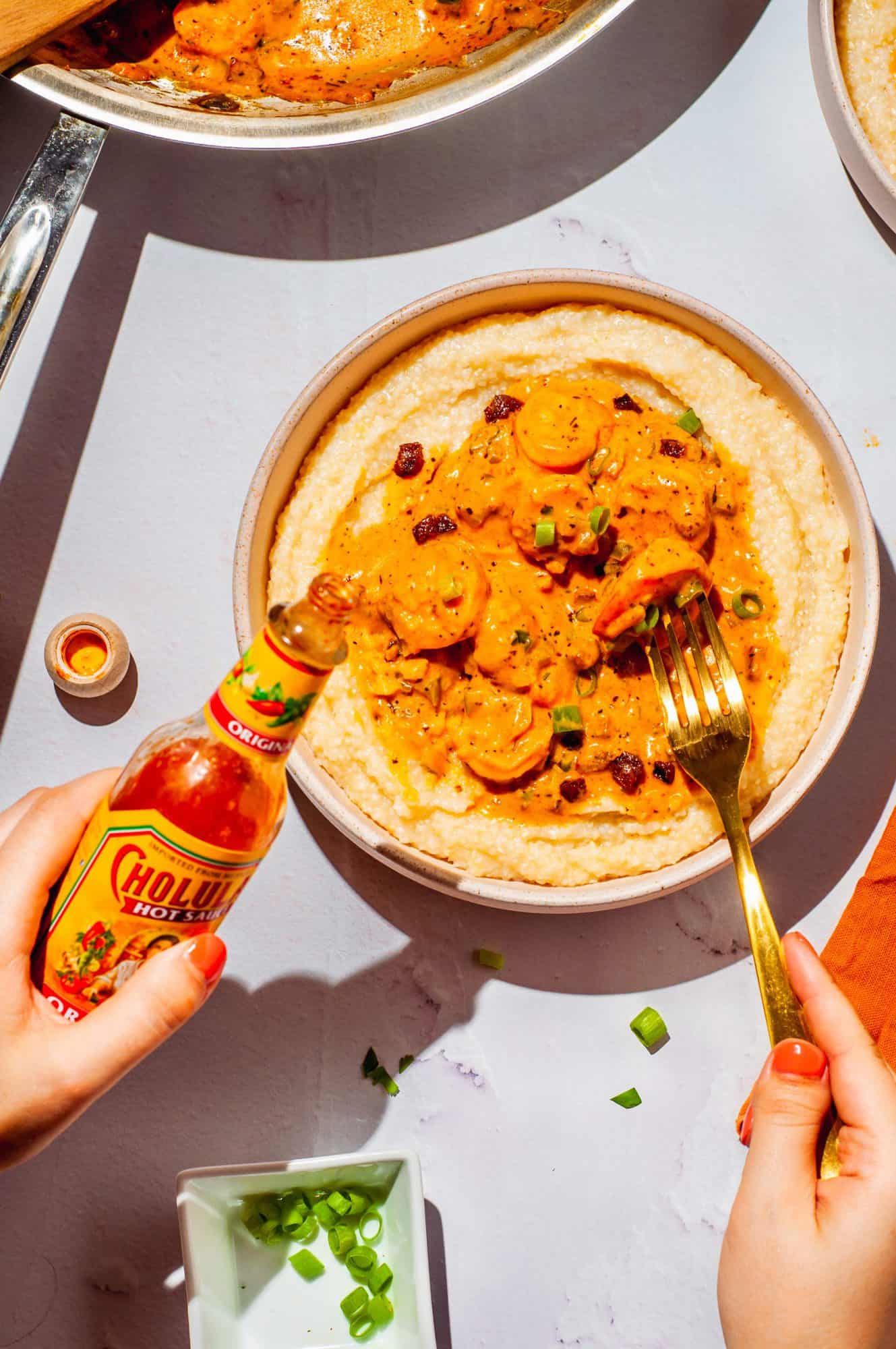 bowl with shrimp and grits with cajun sauce, hands holding fork, bottle of hot sauce