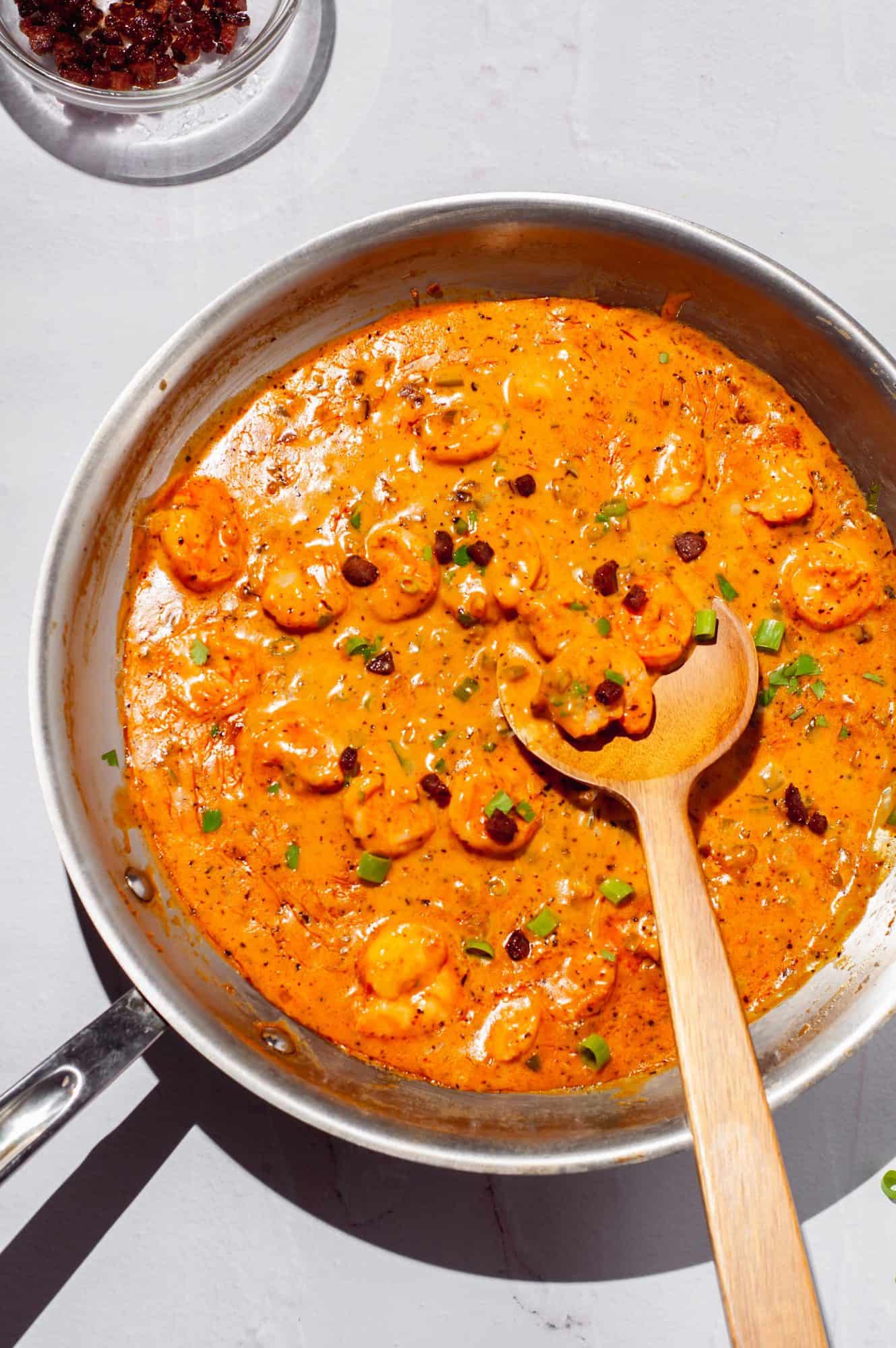 Shrimp tossed in Cajun cream sauce in a pan with spoon, topped with Andouille sausage and scallions