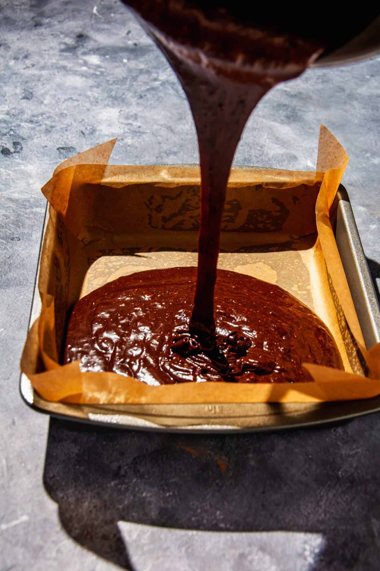action shot shows pouring brownie batter from a bowl into a square baking pan lined with parchment paper