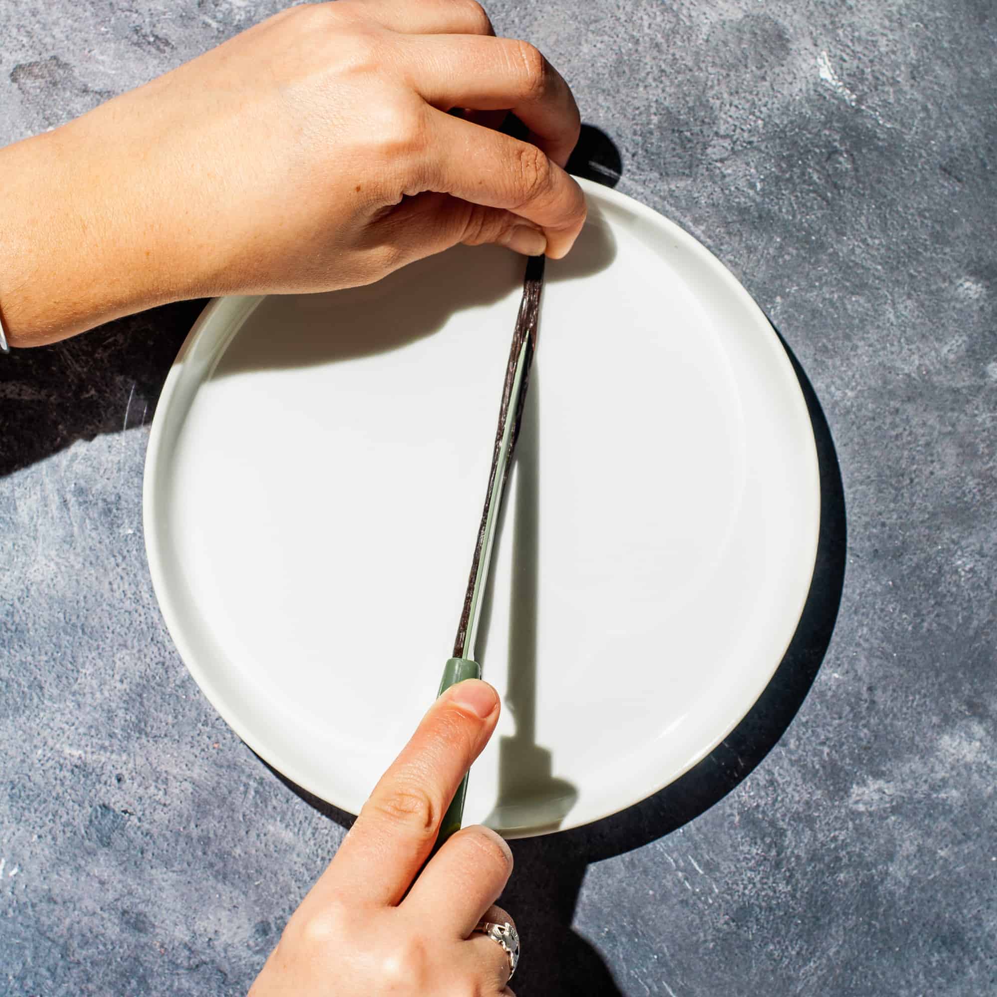 Hands slicing a vanilla bean lengthwise down the center with a paring knife on a white plate