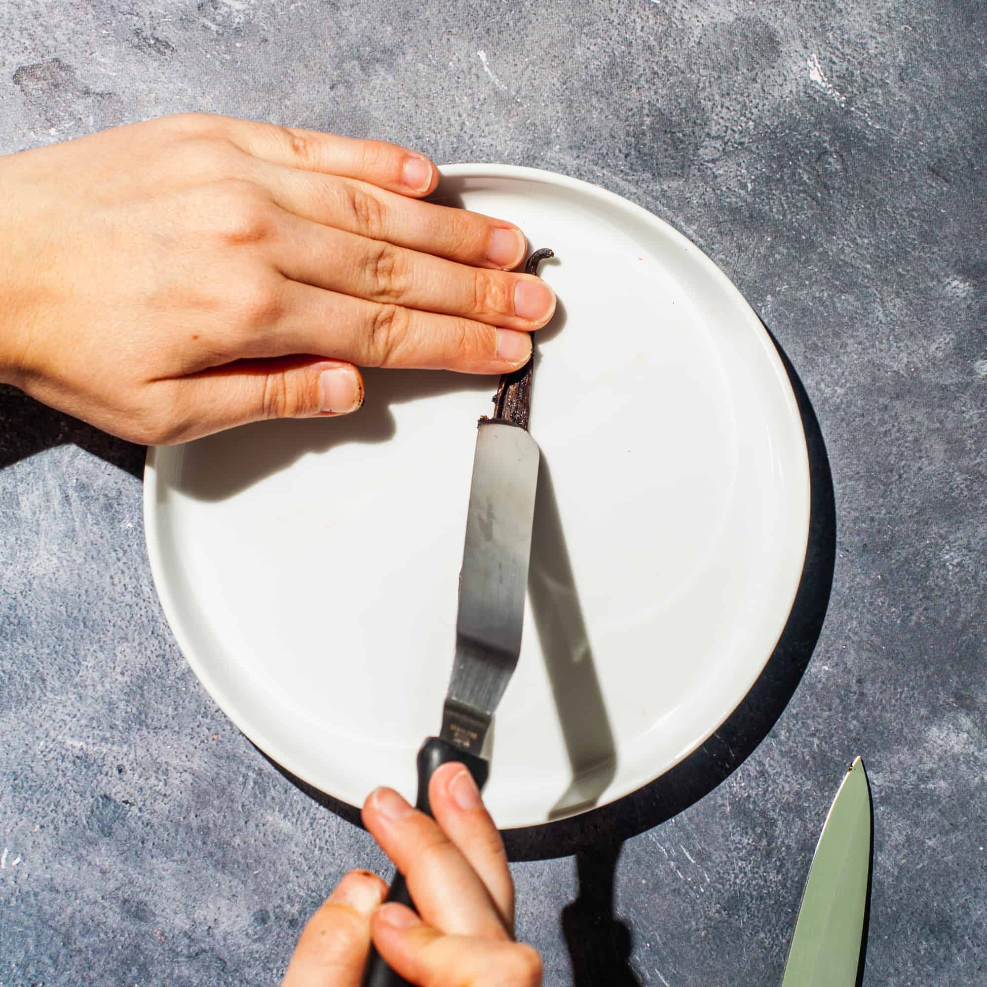 Hands using an offset spatula to scrape seeds from the inside of a vanilla bean