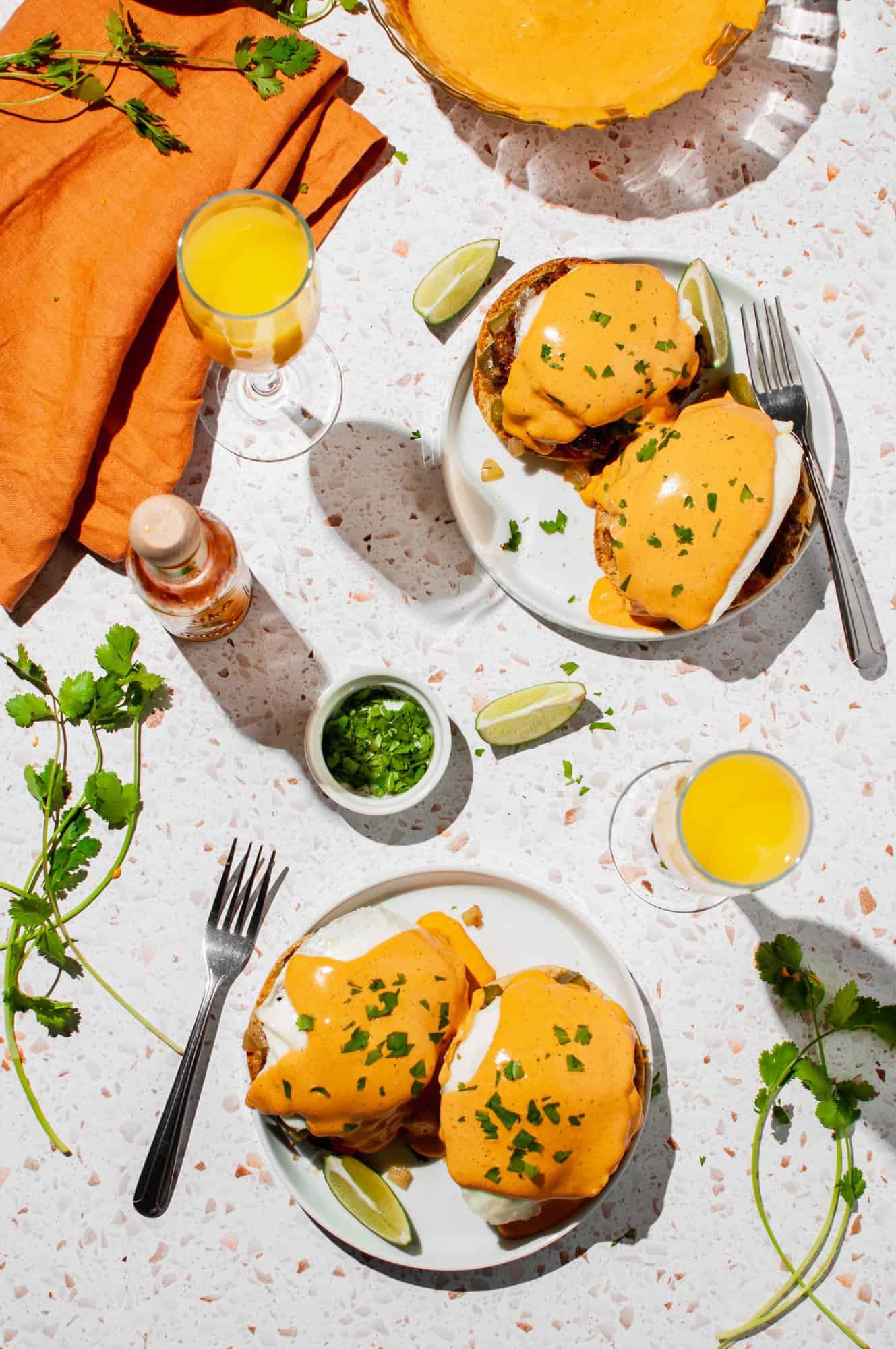 two plates with eggs benedict with chipotle hollandaise, cilantro, two mimosas, two forks, lime wedges, hot sauce