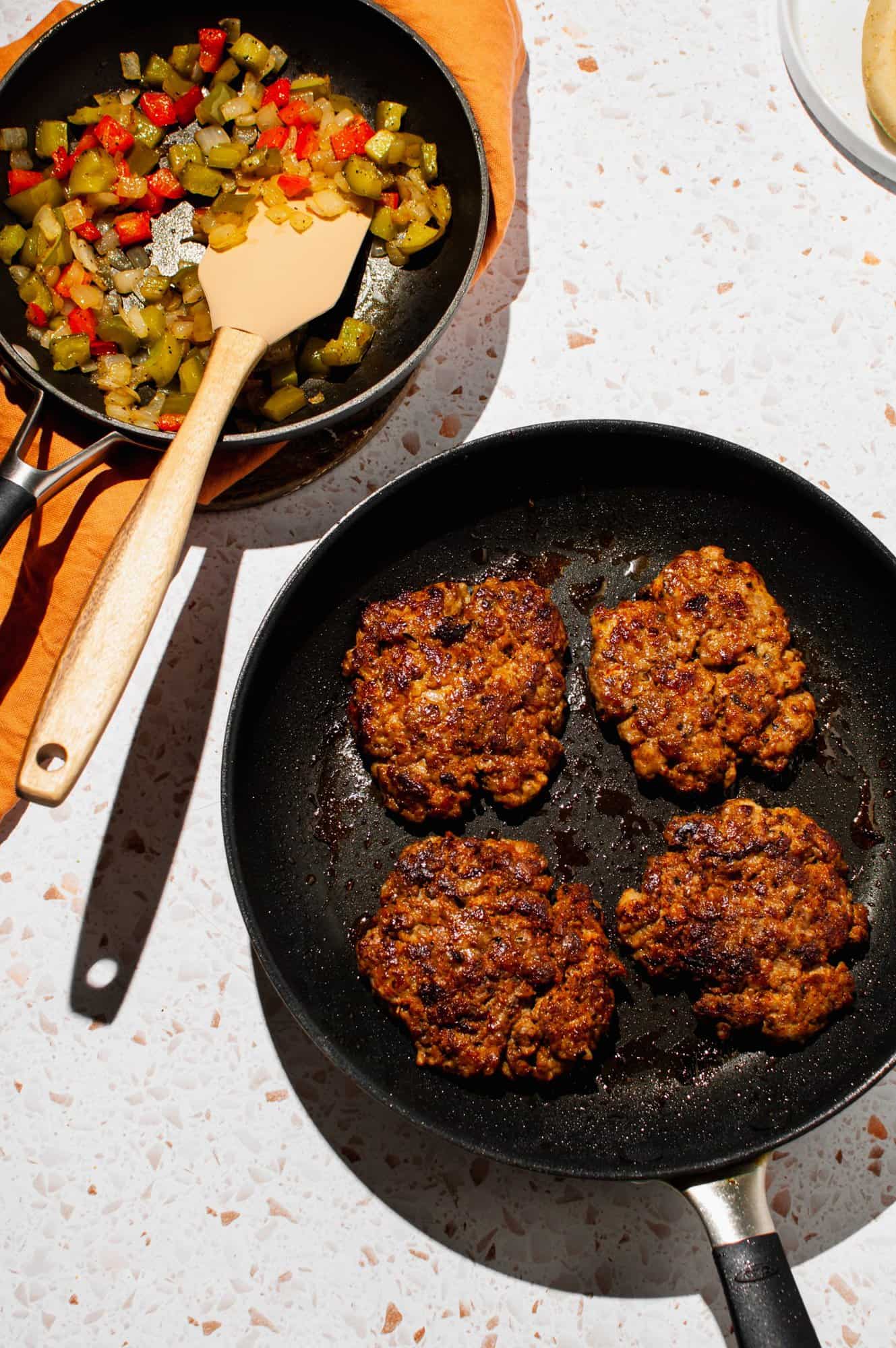 4 chorizo patties cooking in a pan, peppers and onions on the side