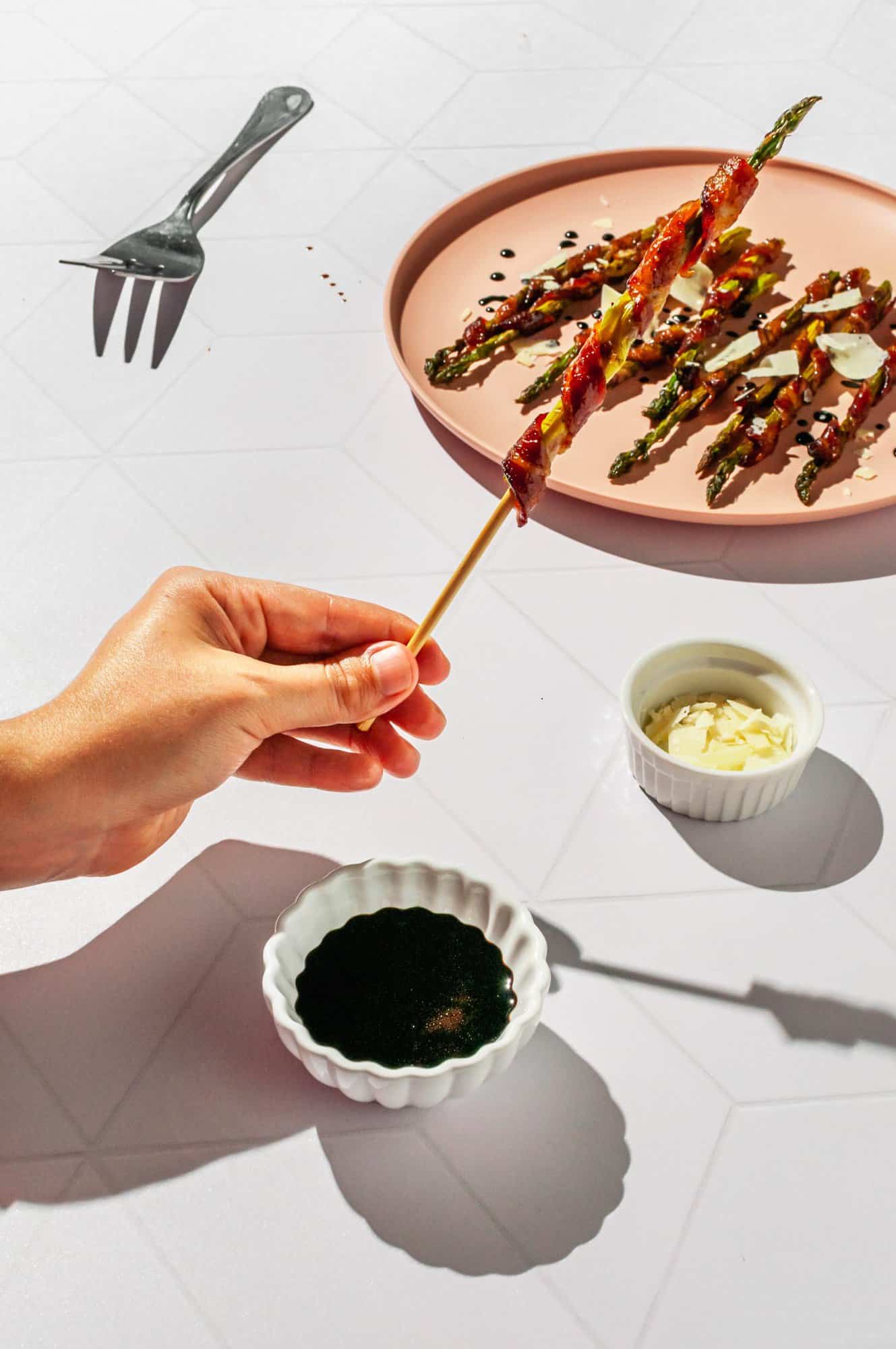 hand holding a bacon wrapped asparagus skewer over a plate of more skewers