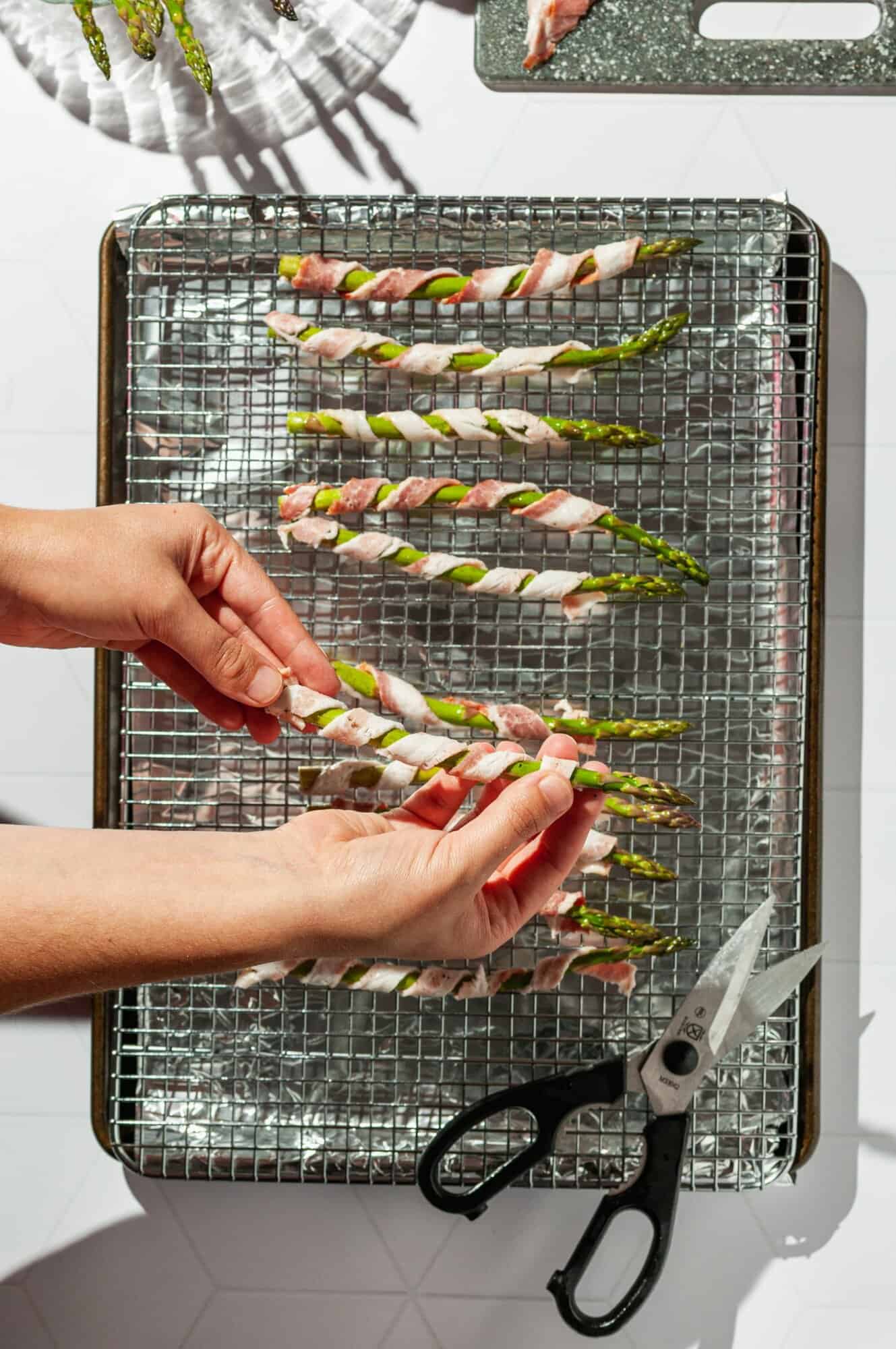 hands holding a piece of asparagus wrapped with bacon above a tray with more asparagus