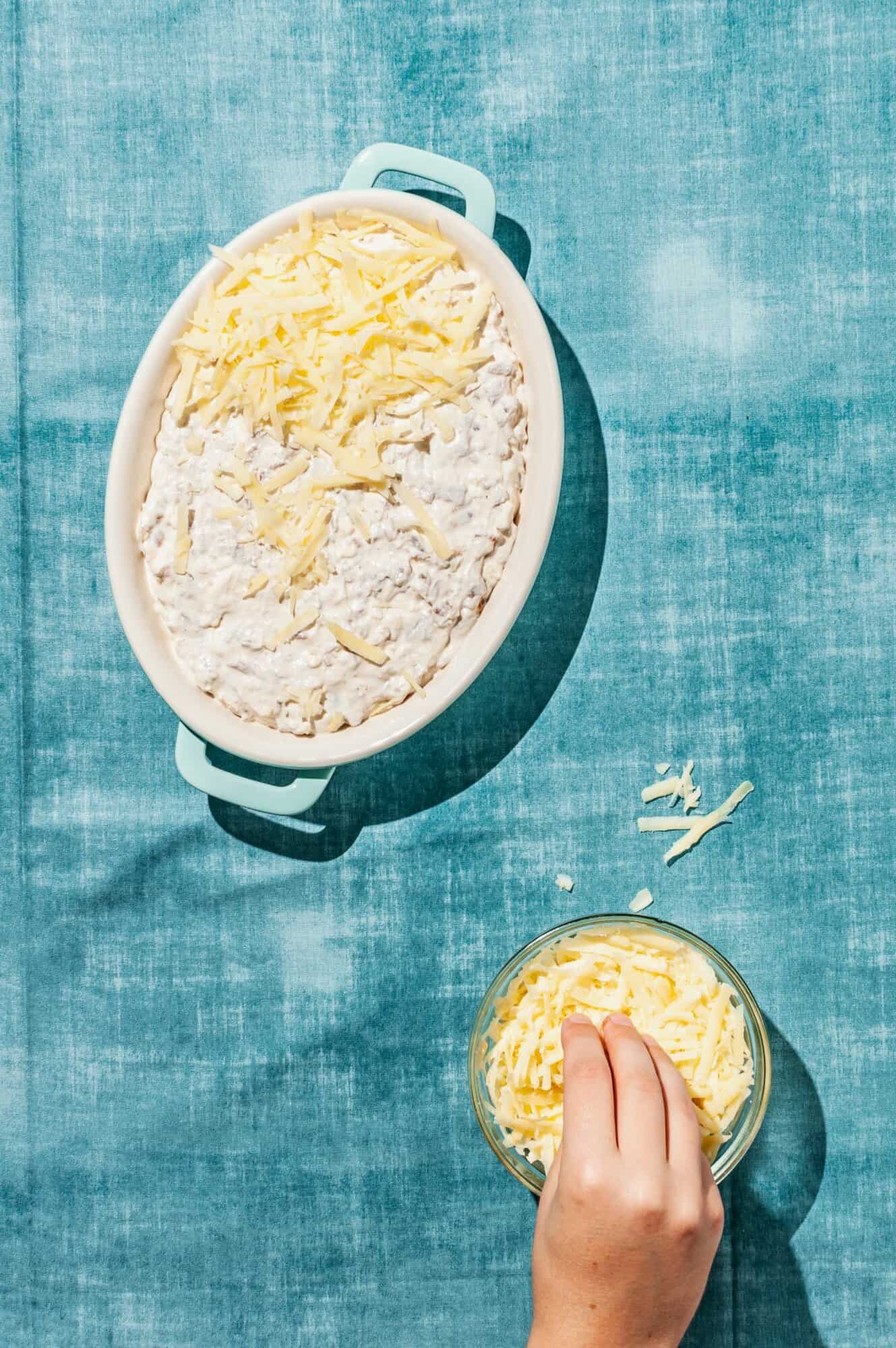 hand reaching into bowl of grated cheese to top caramelized onion dip