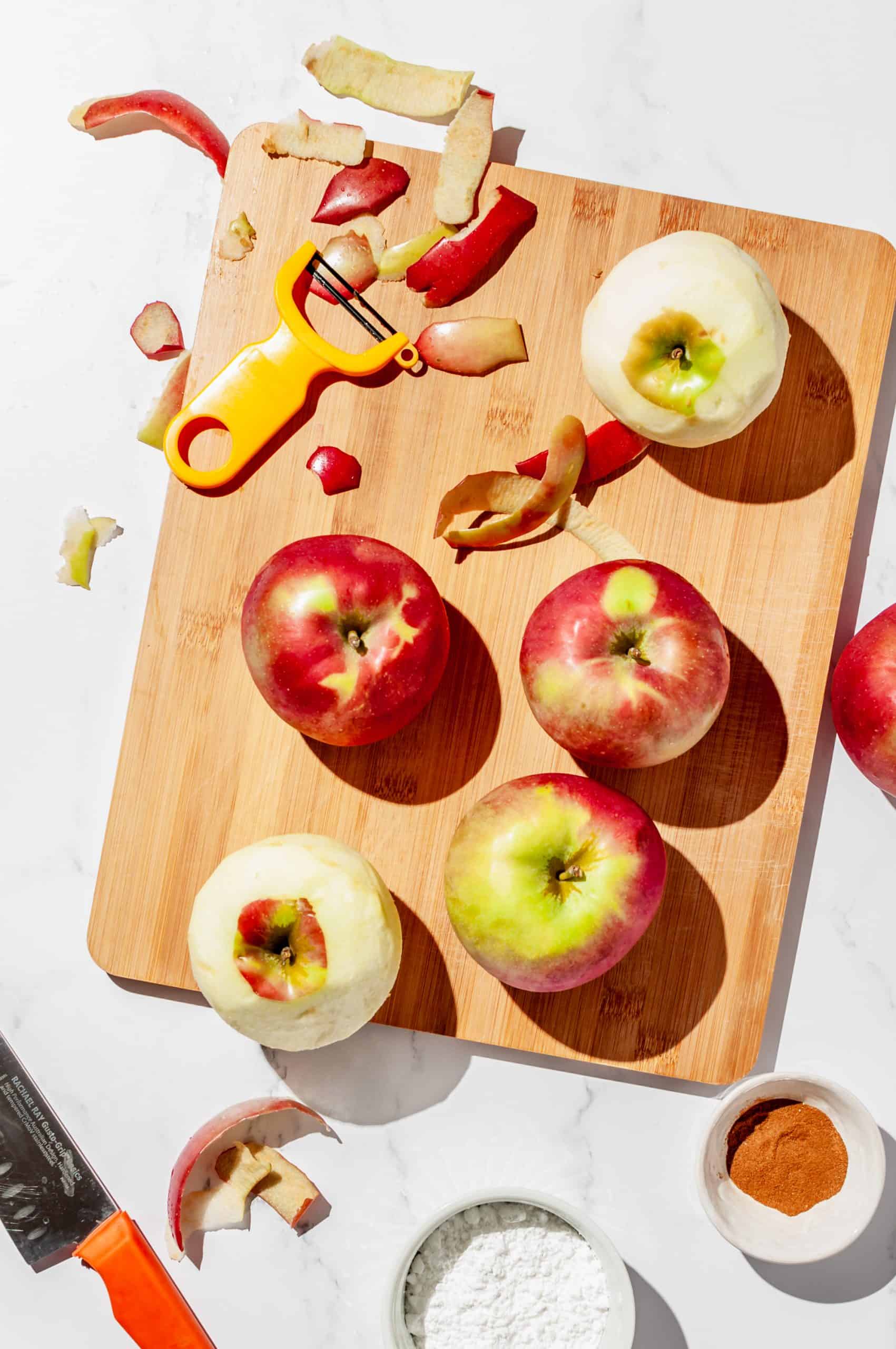5 apples on cutting board, two peeled, with yellow peeler and scraps