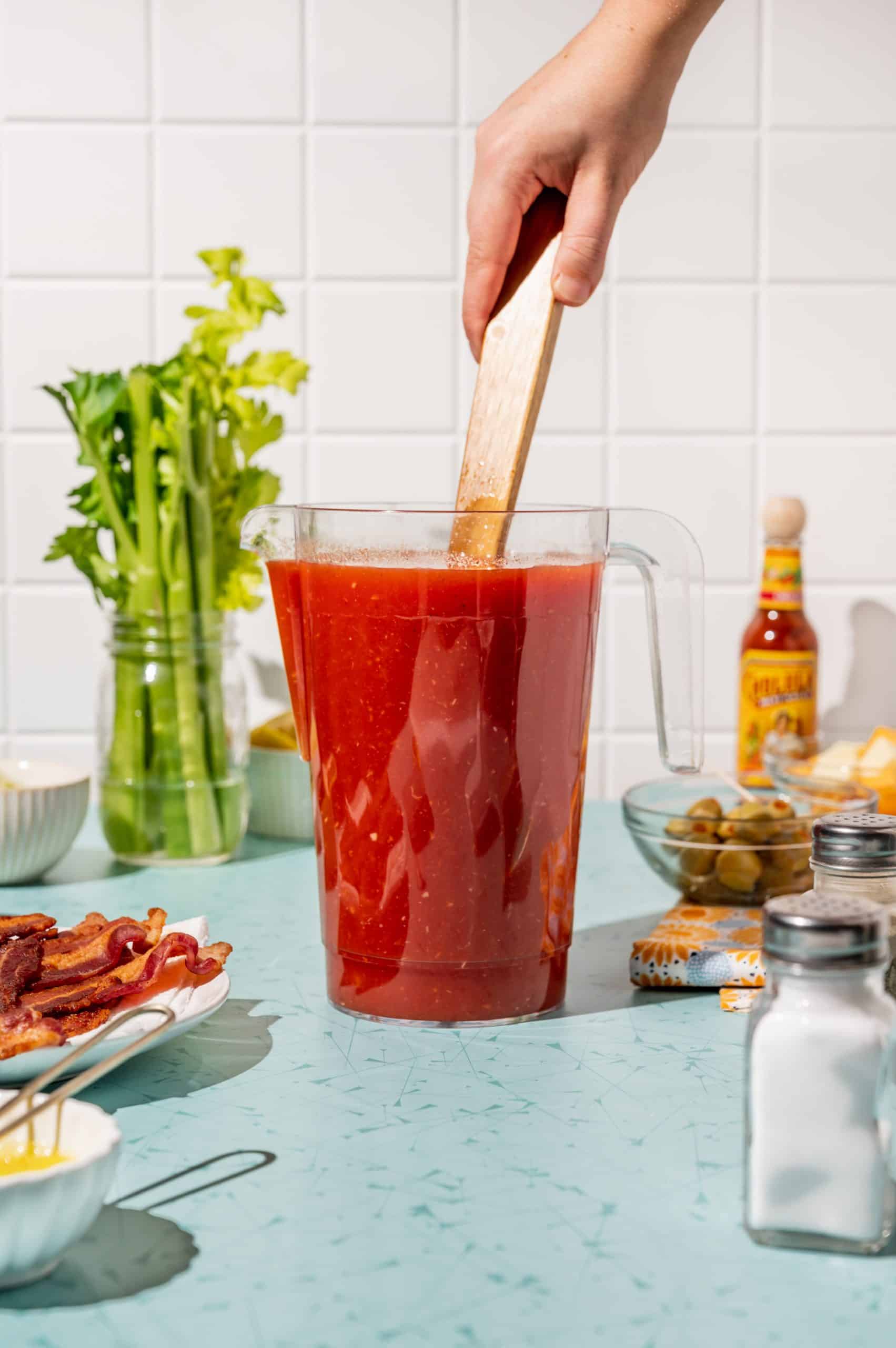 hand stirring bloody mary mix in a large pitcher with a wooden spoon