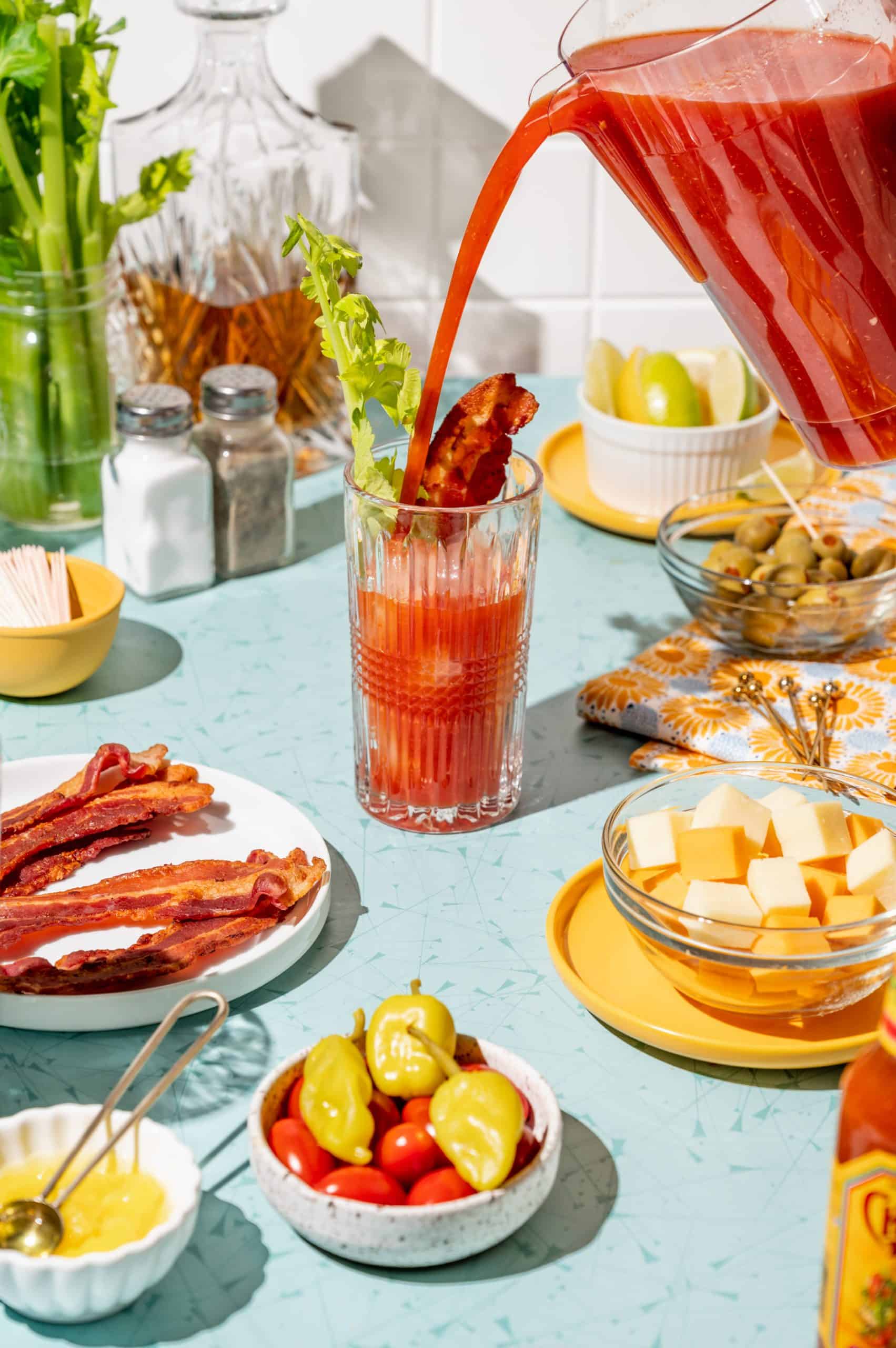 Pouring bloody mary mix into a glass with ice, a strip of bacon and a celery stalk