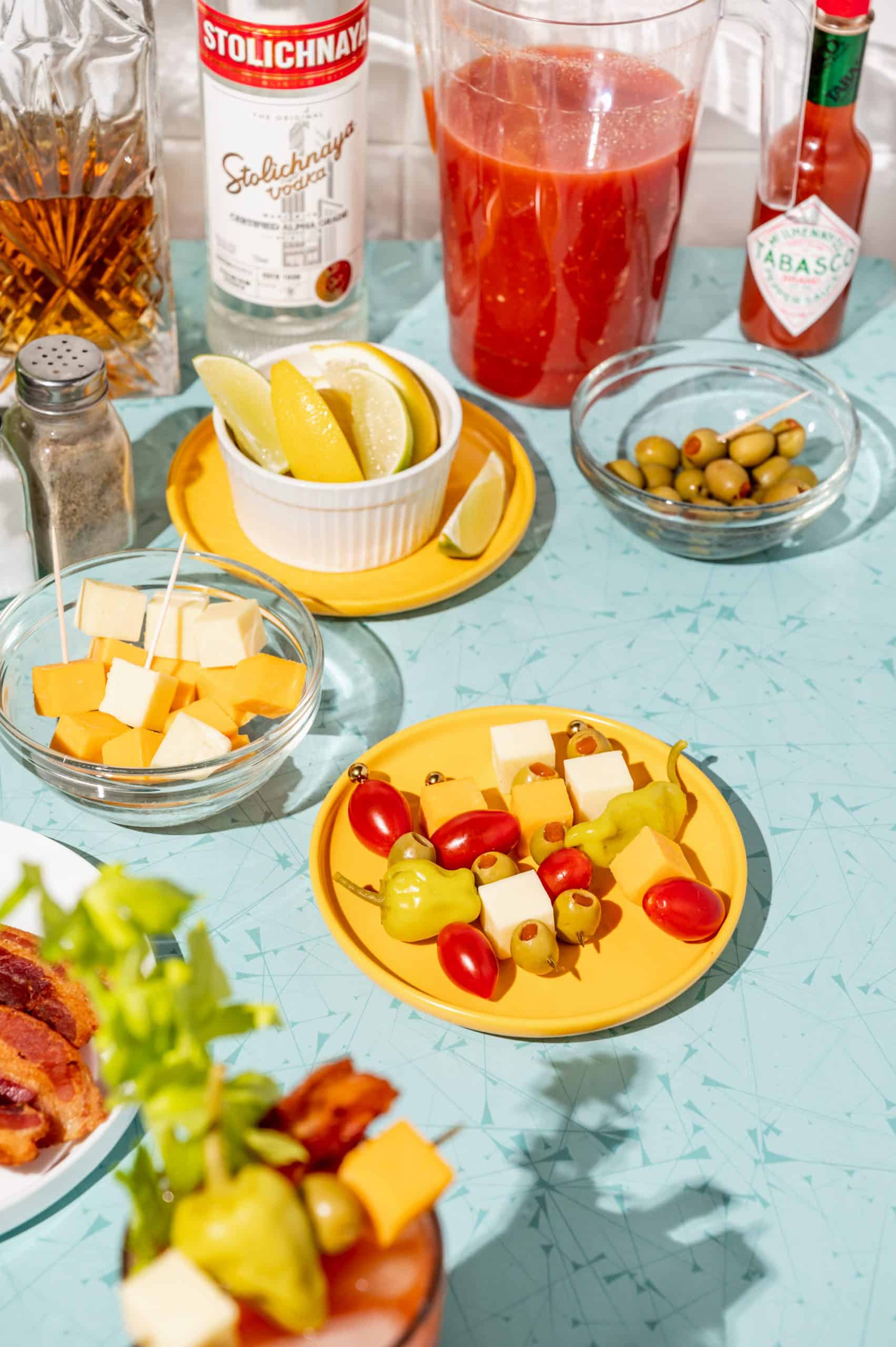 DIY bloody mary bar with a plate of premade cocktail picks with olives, peppers, cheese cubes, and cherry tomatoes