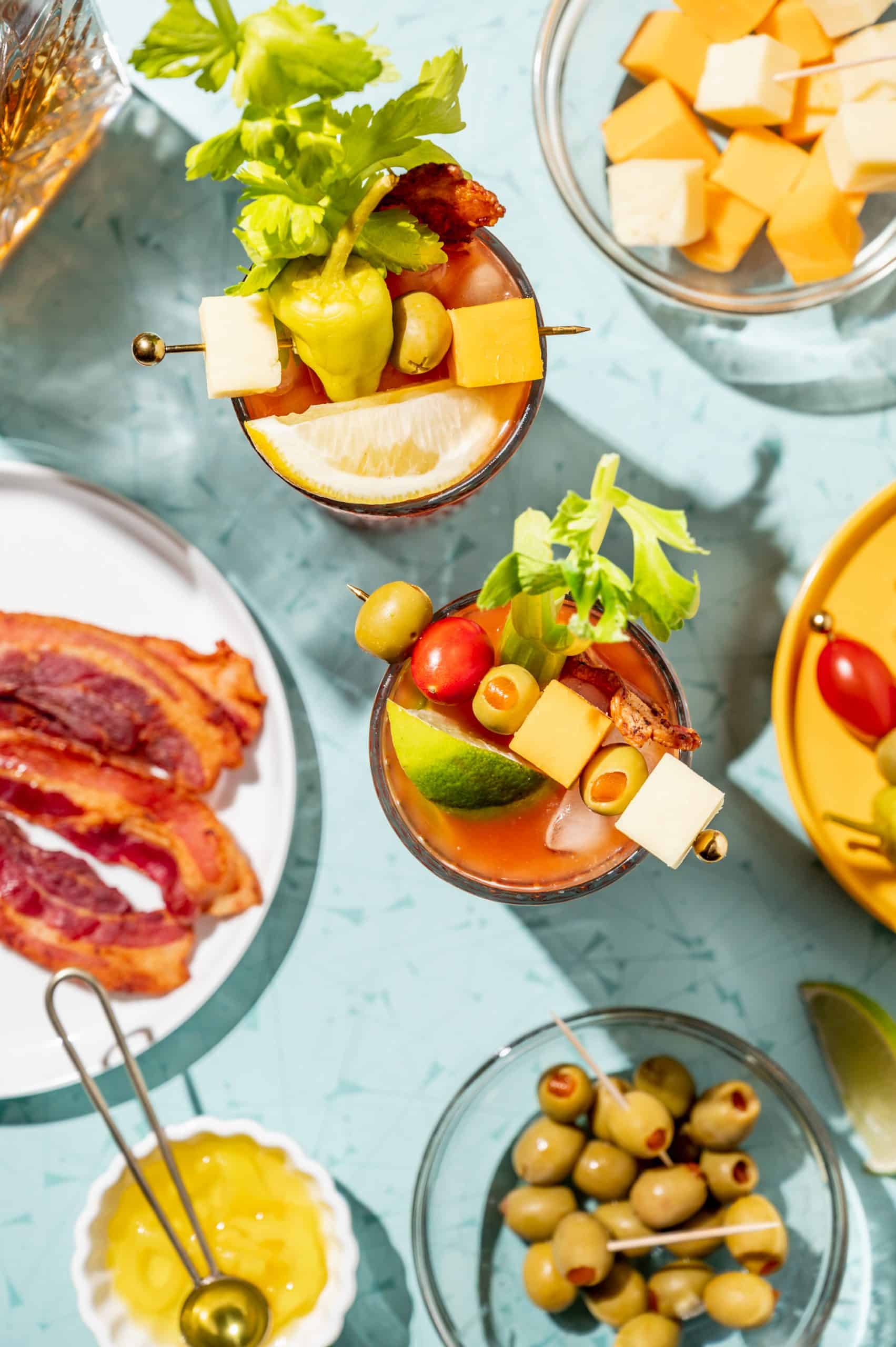 two bloody marys with garnishes on top: bacon, celery, lime wedge, olives, cheese cubes