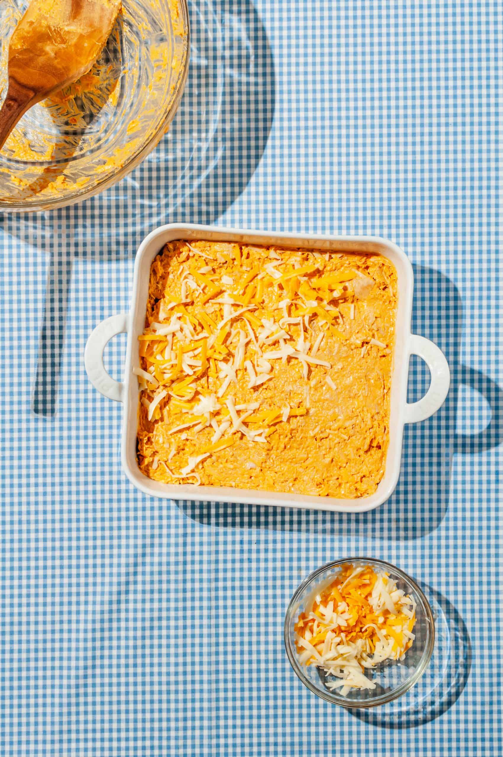 buffalo chicken dip mixture in a baking dish topped with shredded cheddar cheese