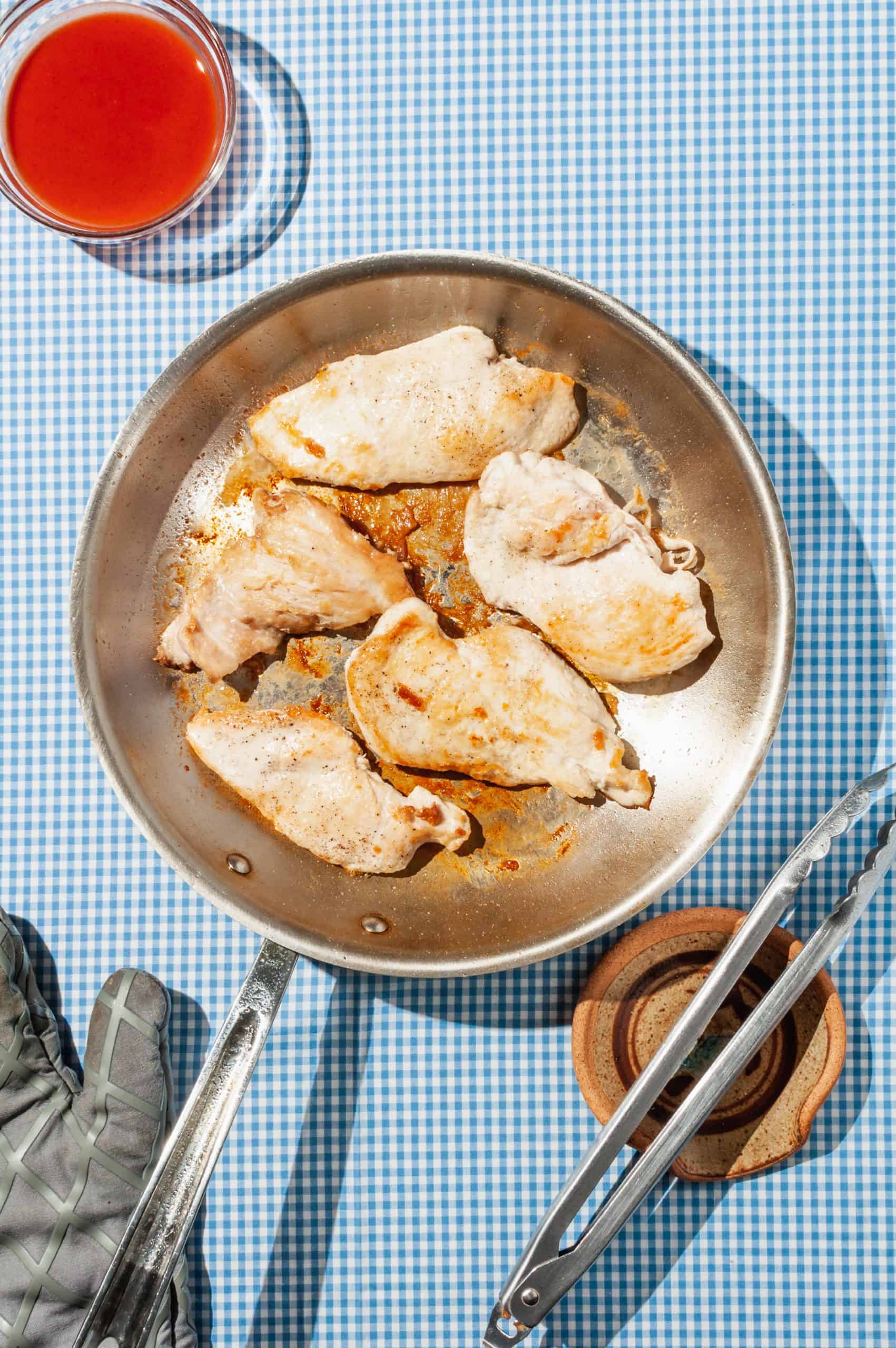 chicken pieces browned in a skillet with a bowl of hot sauce on the side