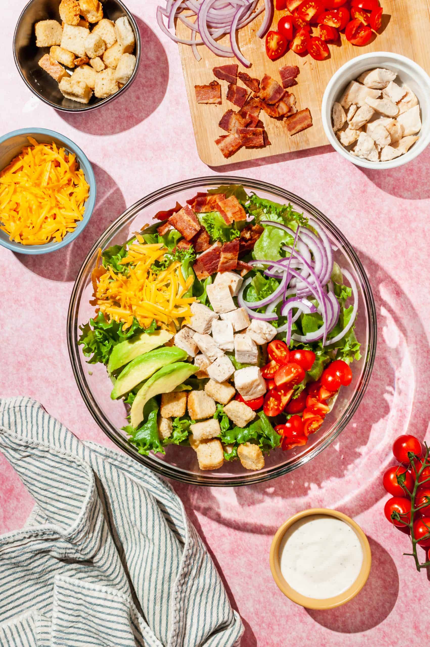 large glass bowl with lettuce, bacon bits, chicken, tomatoes, sliced red onion, croutons, cheddar, before mixing