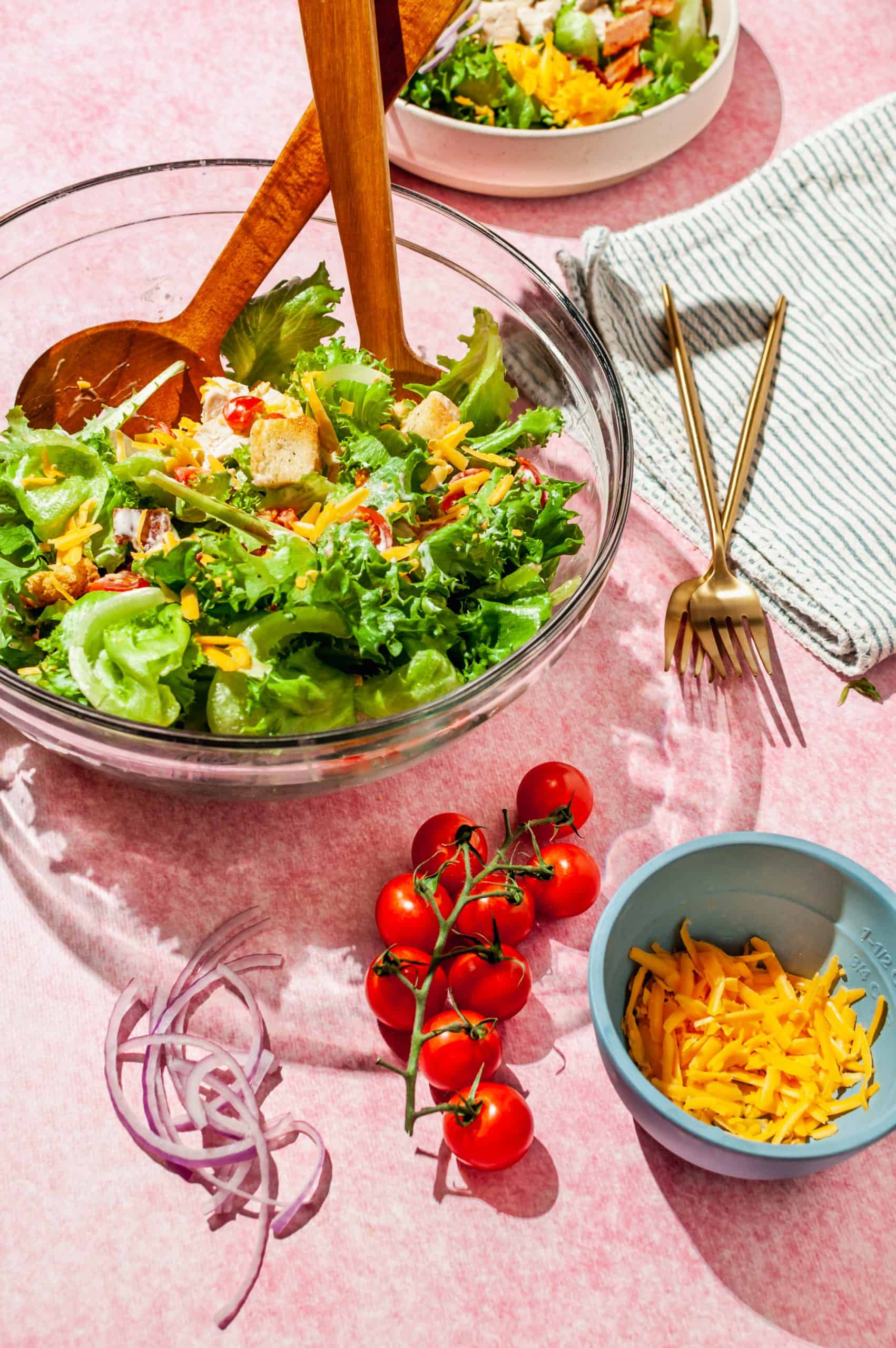 tossing salad in a large glass bowl with extra onion, tomato and cheese in front of bowl