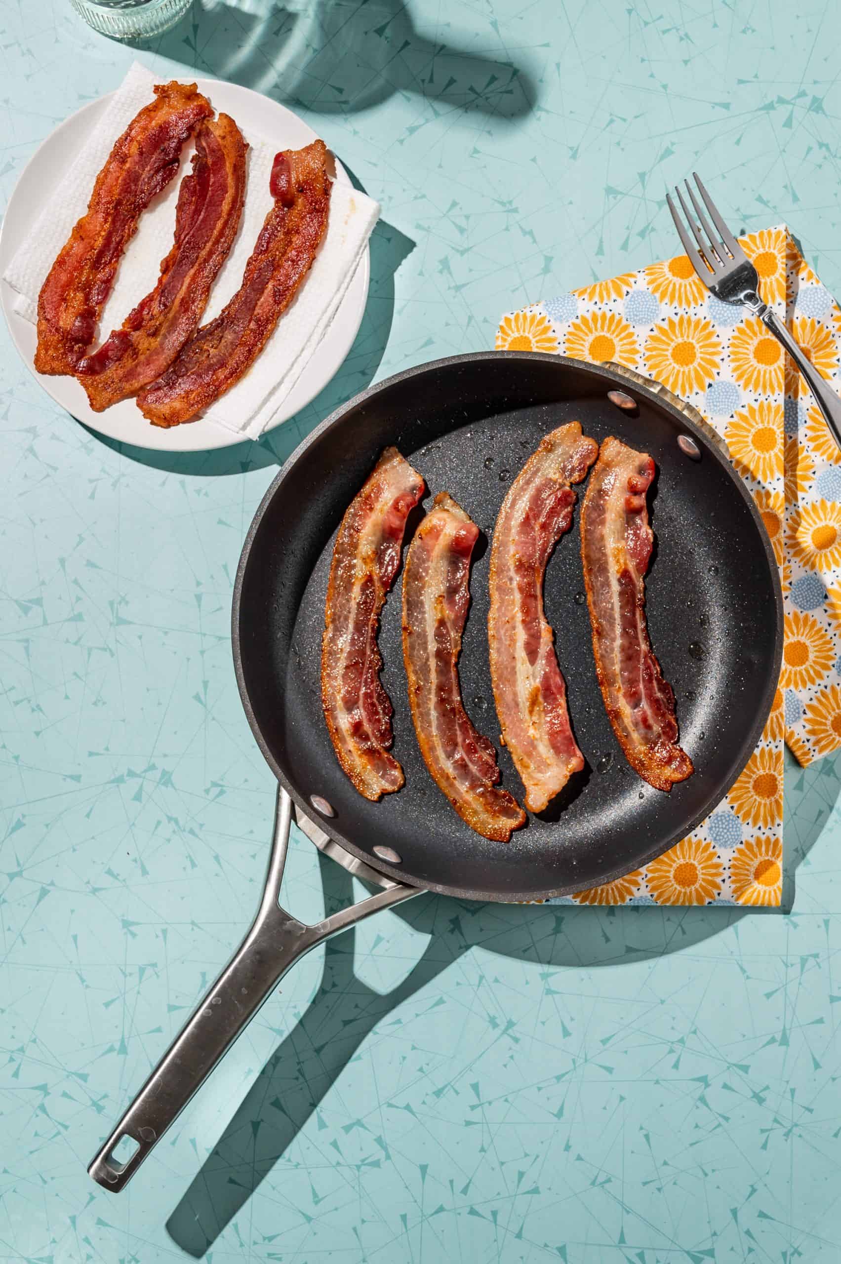4 strips of bacon pan frying in a skillet, plate of cooked bacon on the side