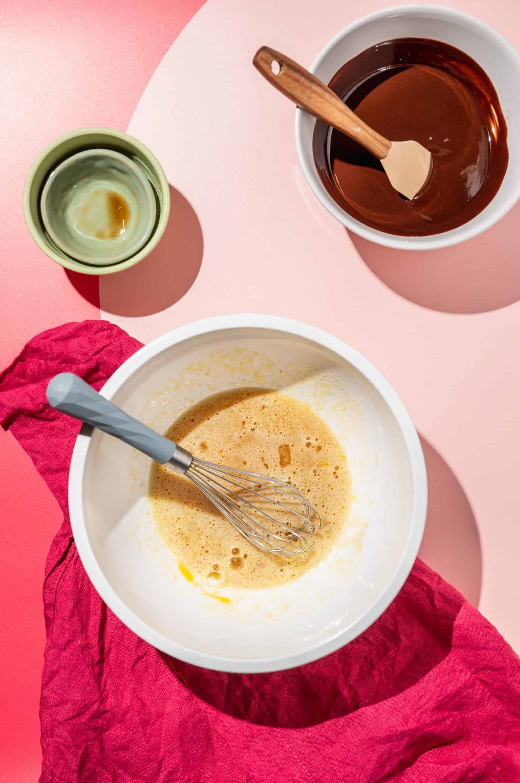 large bowl with egg mixture with a whisk in it, bowl of melted chocolate to the side