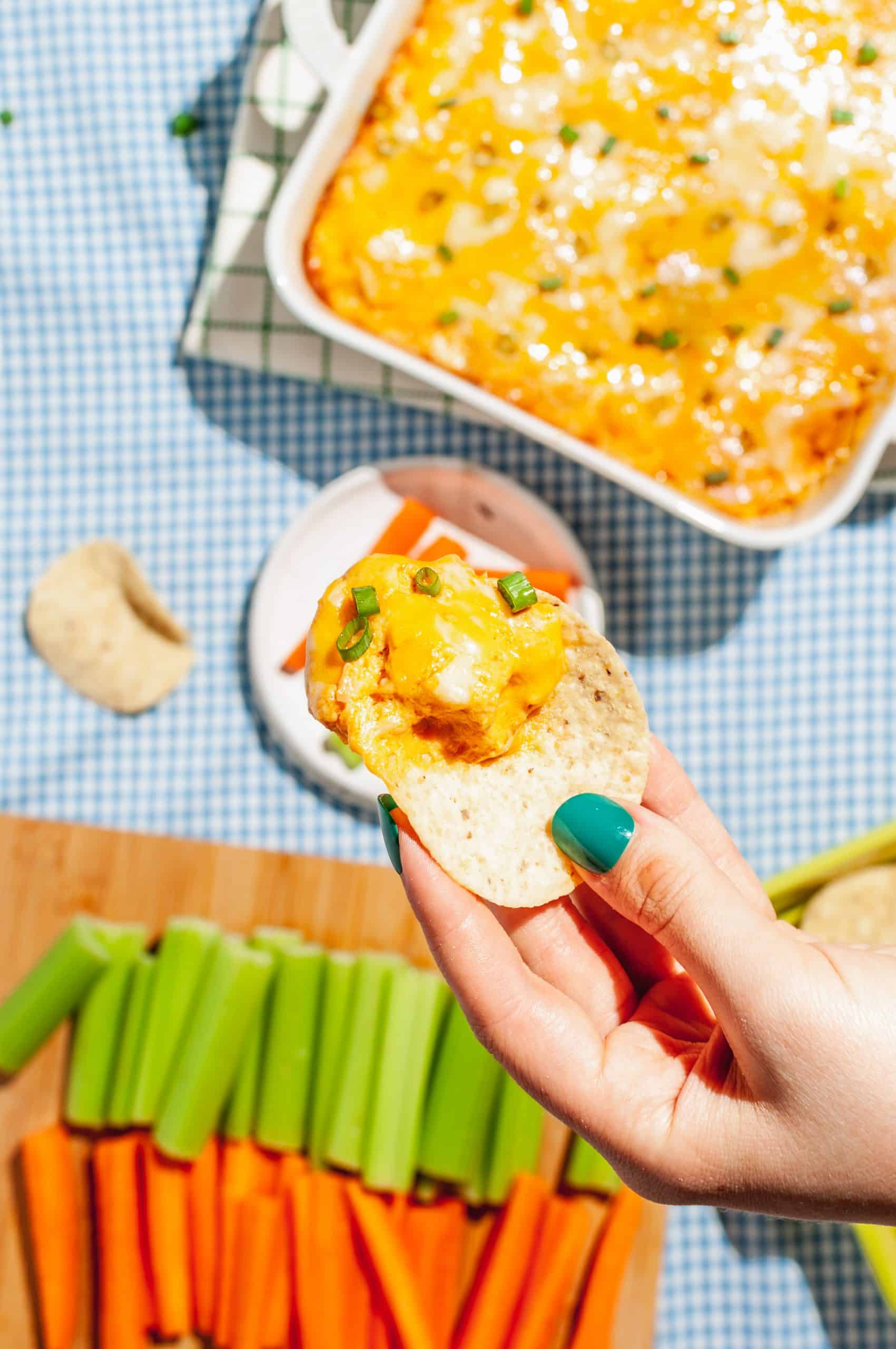 hand holding a tortilla chip with buffalo chicken dip above tray of carrots and celery