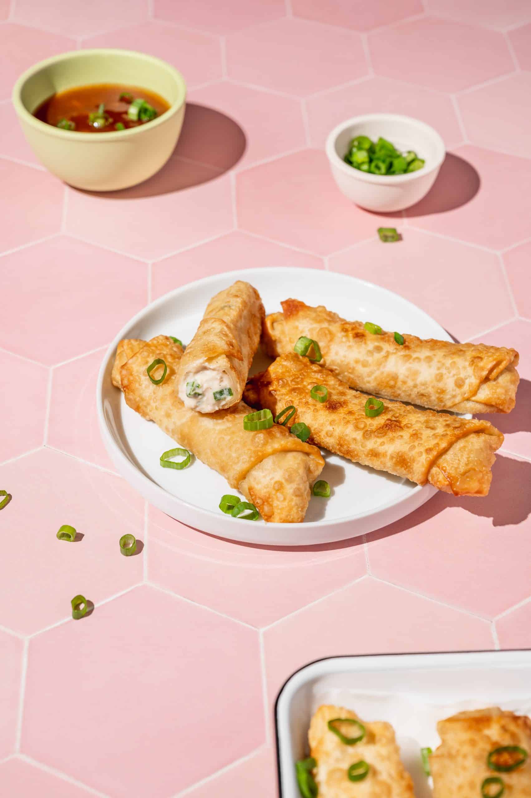 4 egg rolls on a white plate garnished with scallions, top one is sliced open to show crab rangoon filling
