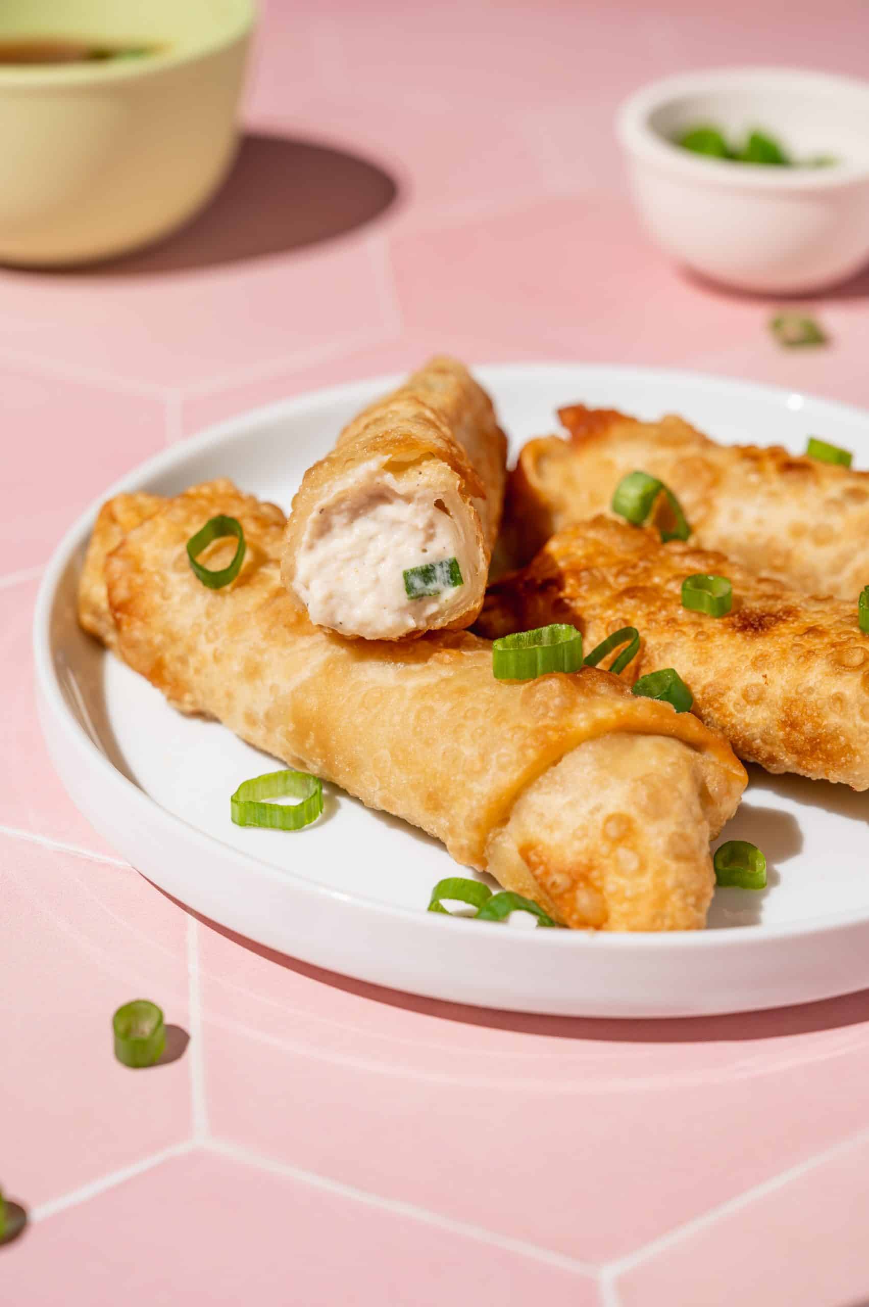 4 egg rolls on a white plate garnished with scallions, top one is sliced open to show crab rangoon filling