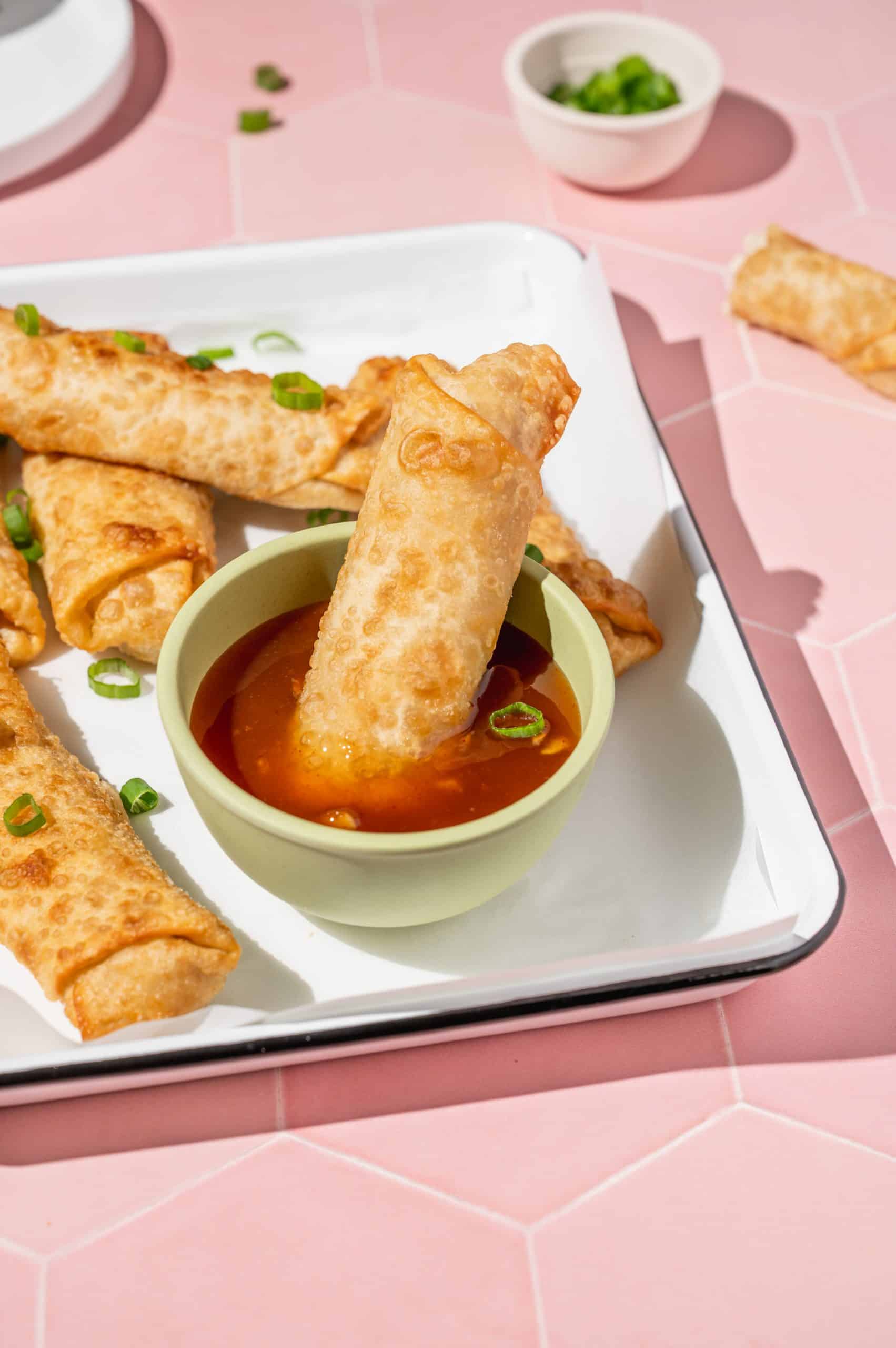 crab rangoon egg roll being dipped in sweet chili sauce, more egg rolls on the tray in the background