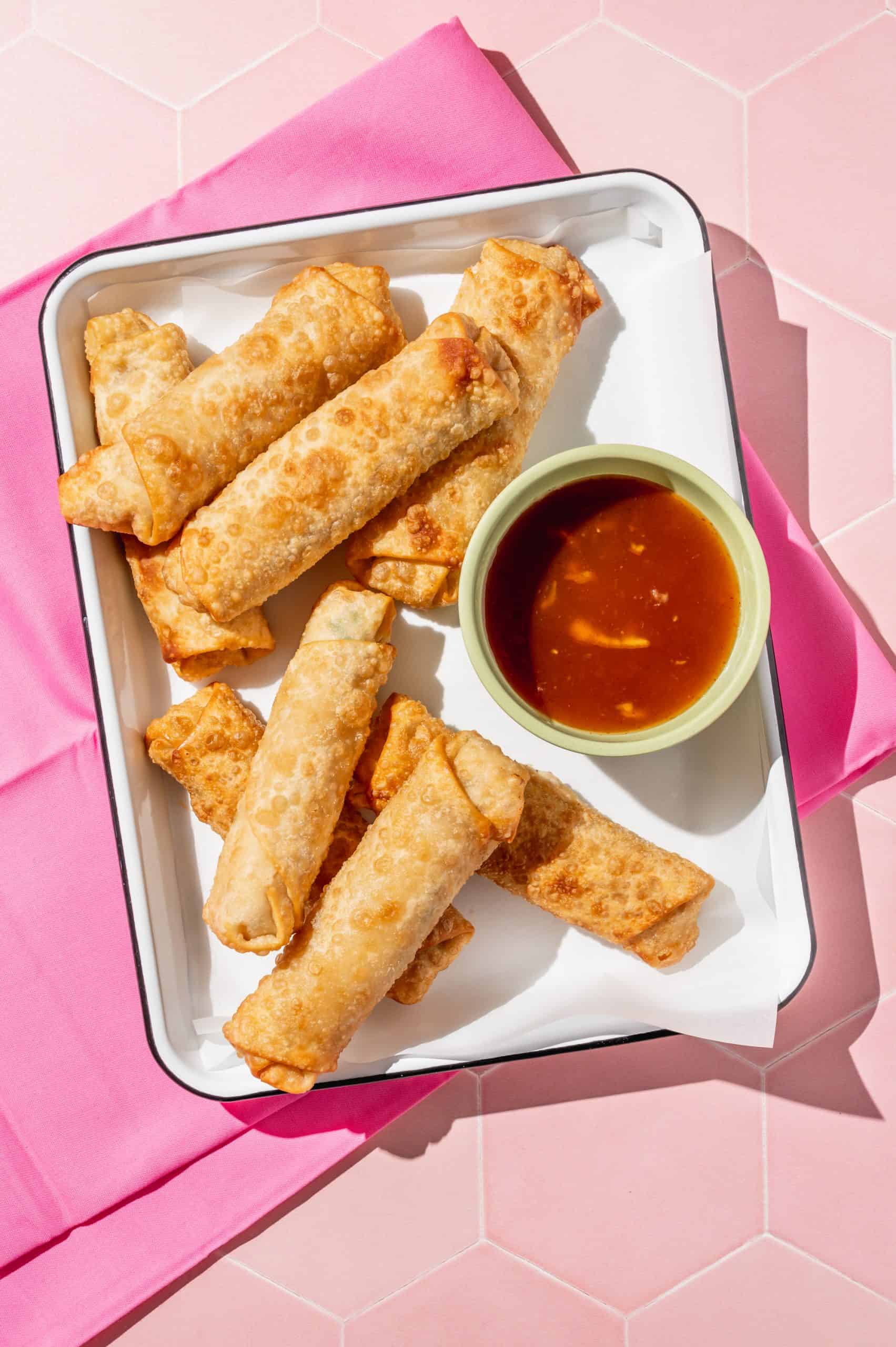 8 fried crab rangoon egg rolls on a tray with cup of sweet chili dipping sauce