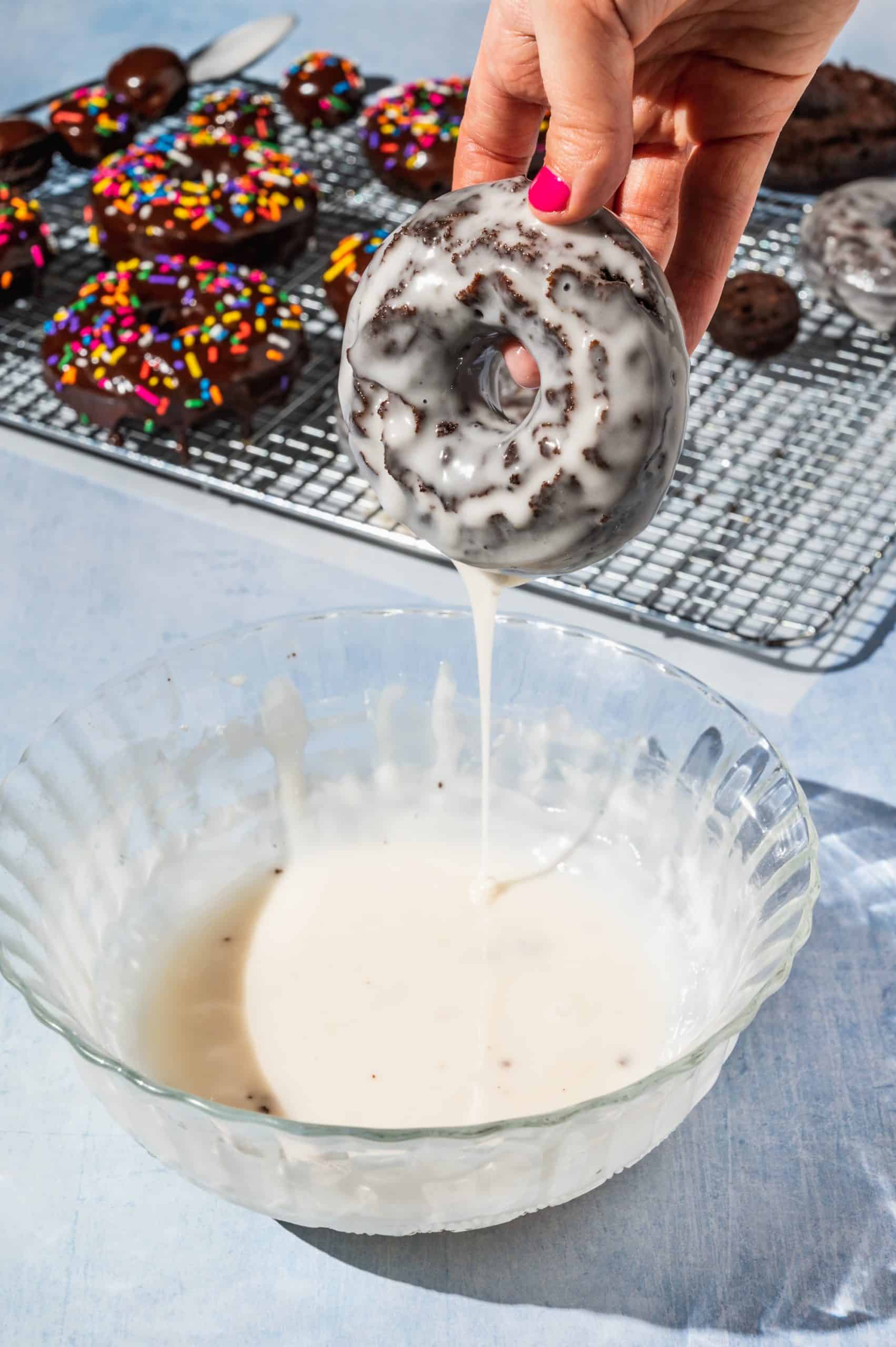 hand holding a chocolate donut above bowl of white glaze after dipping with excess glaze dripping off