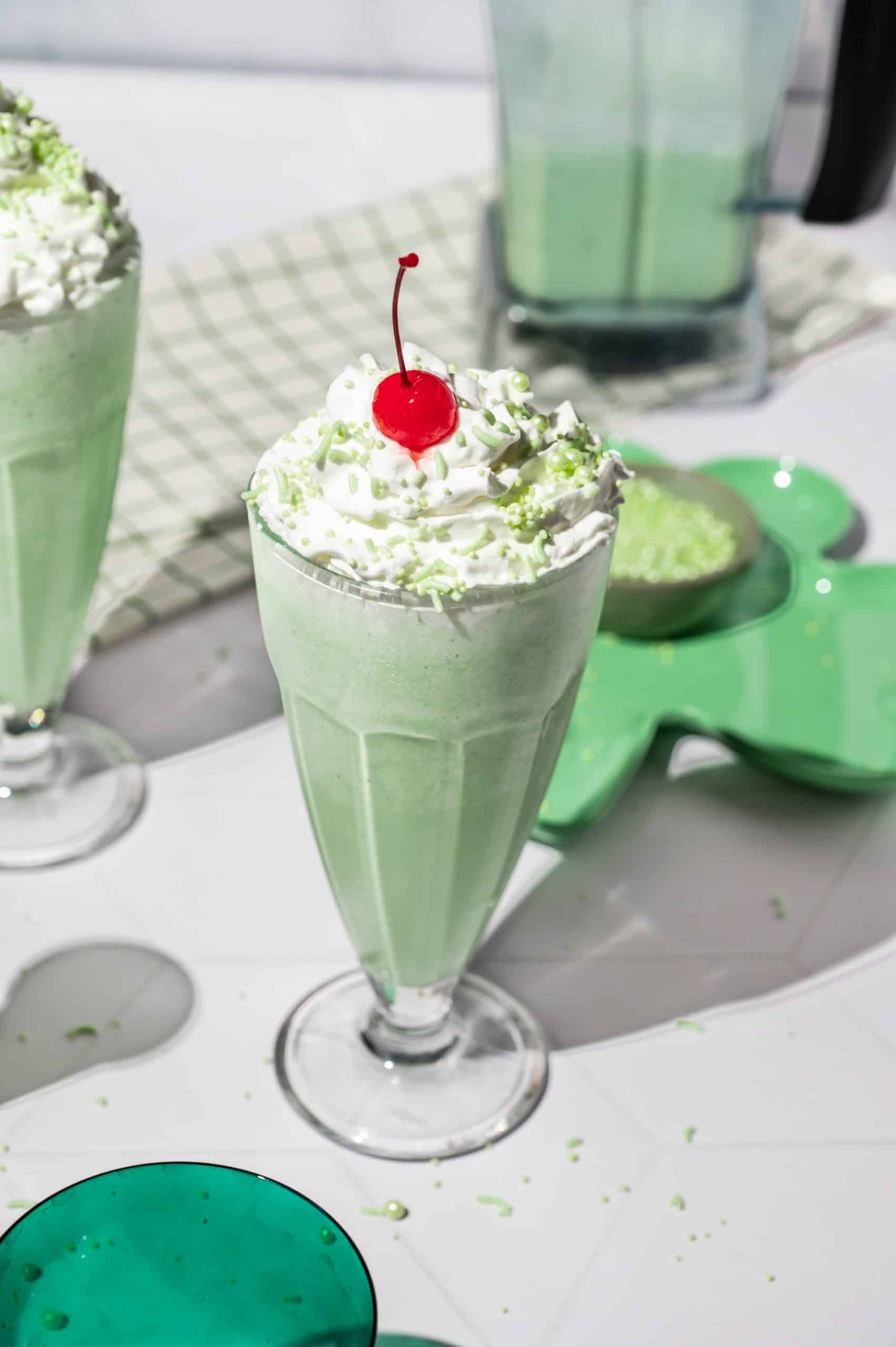 A shamrock shake in a milkshake glass with whipped cream, green sprinkles, and a cherry on top