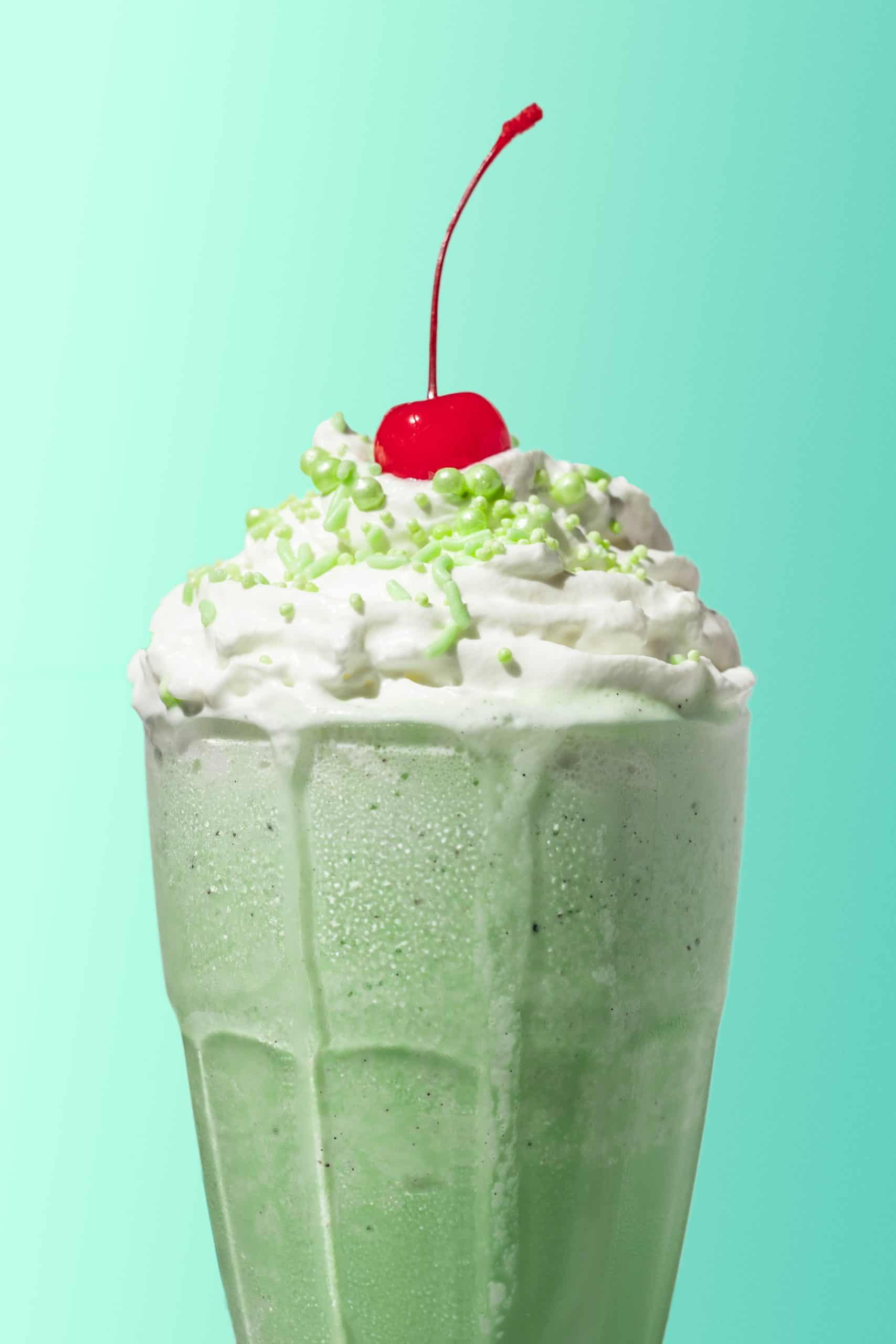 A shamrock shake in a milkshake glass with whipped cream, green sprinkles, and a cherry on top