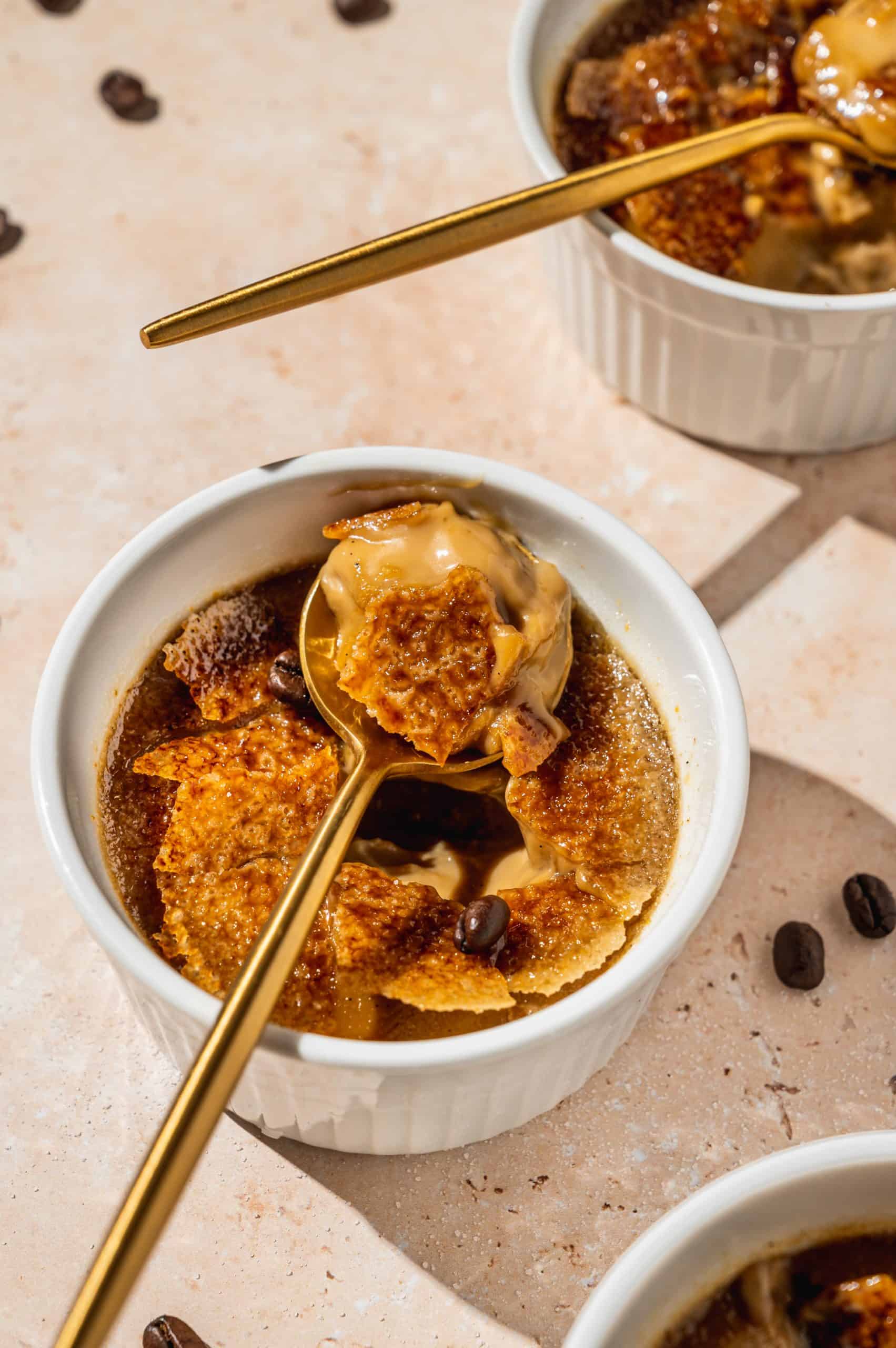 close up of ramekin with coffee creme brulee with a spoon on top showing creamy custard and jagged caramelized sugar pieces