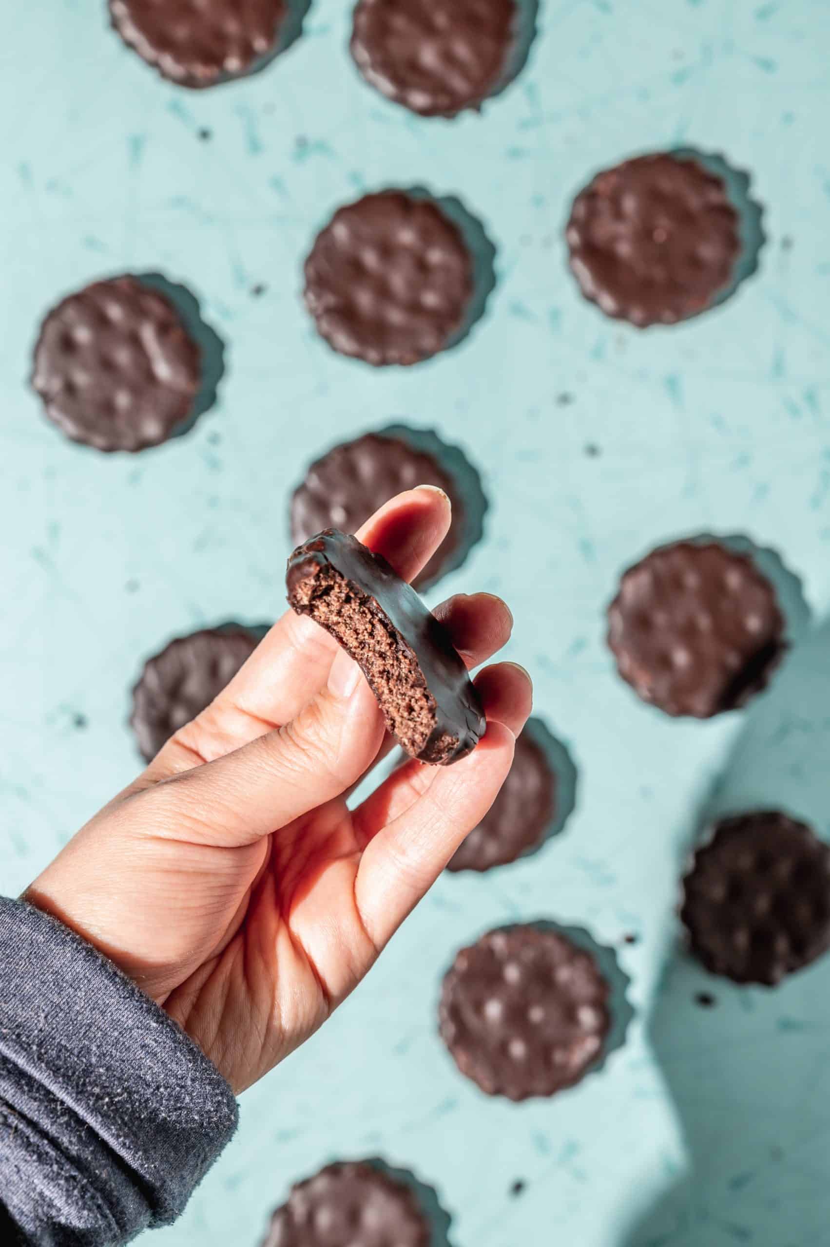 hand holding a thin mint with a bite taken out above more thin mint cookies on a teal speckled background