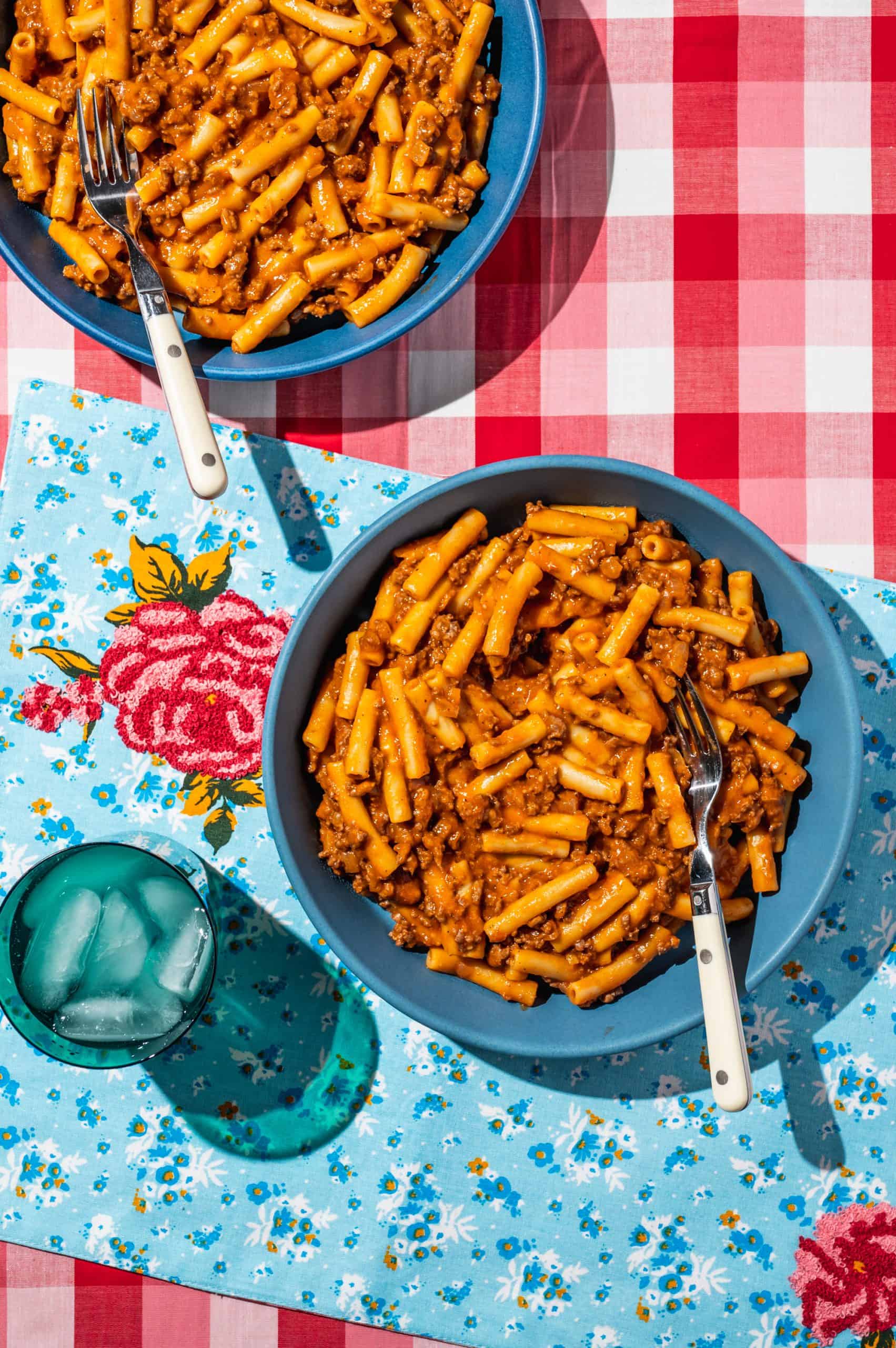 two bowls of homemade beefaroni on a blue placement on a red checkered tablecloth