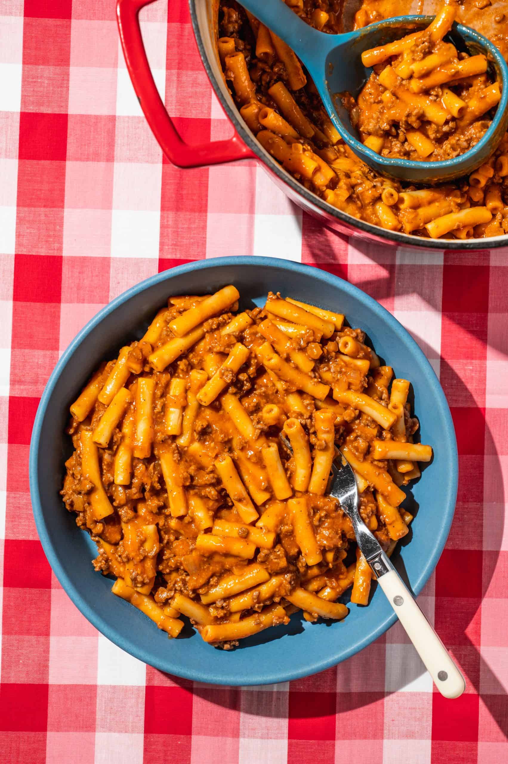 blue shallow bowl with homemade beefaroni, red pot filled with beefroni to the side